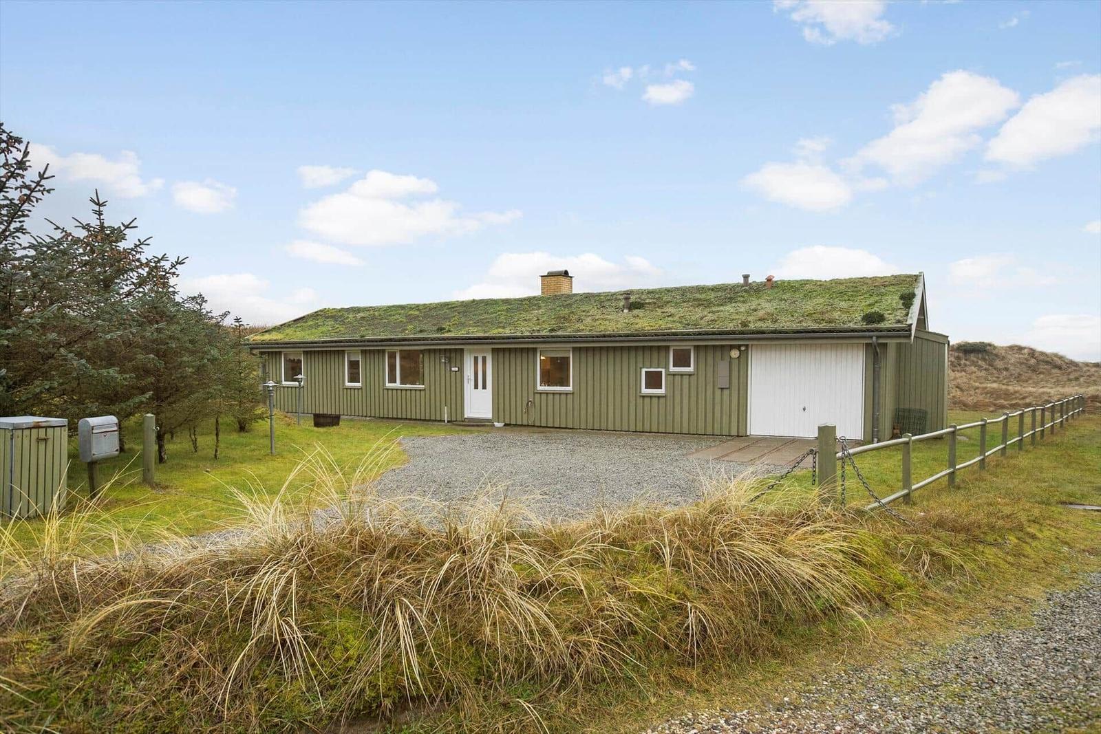 A green house with a green roof and white garage. Surrounded by grass and trees.