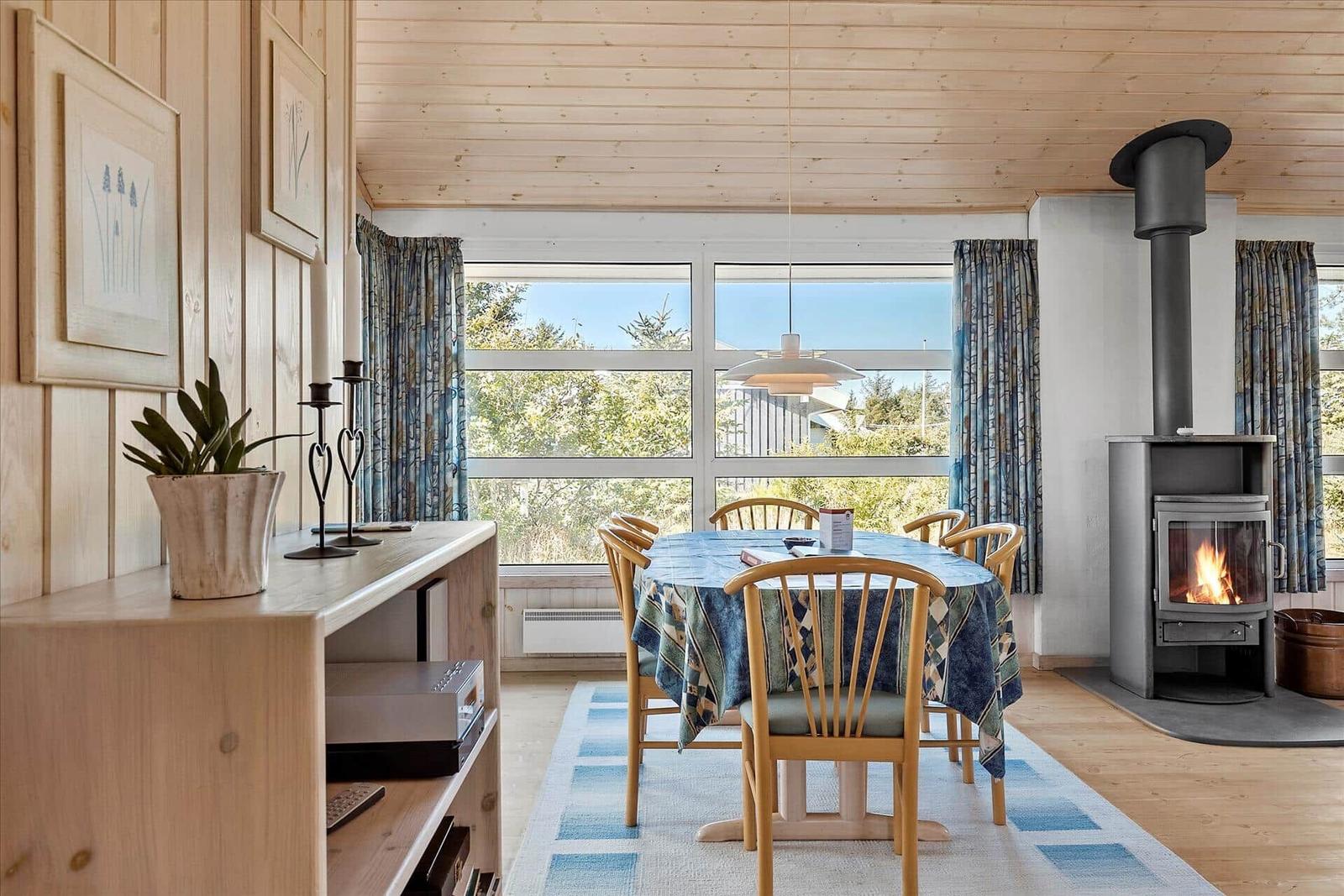 Dining room with wooden table, chairs, and wood stove. Large windows with view of greenery.