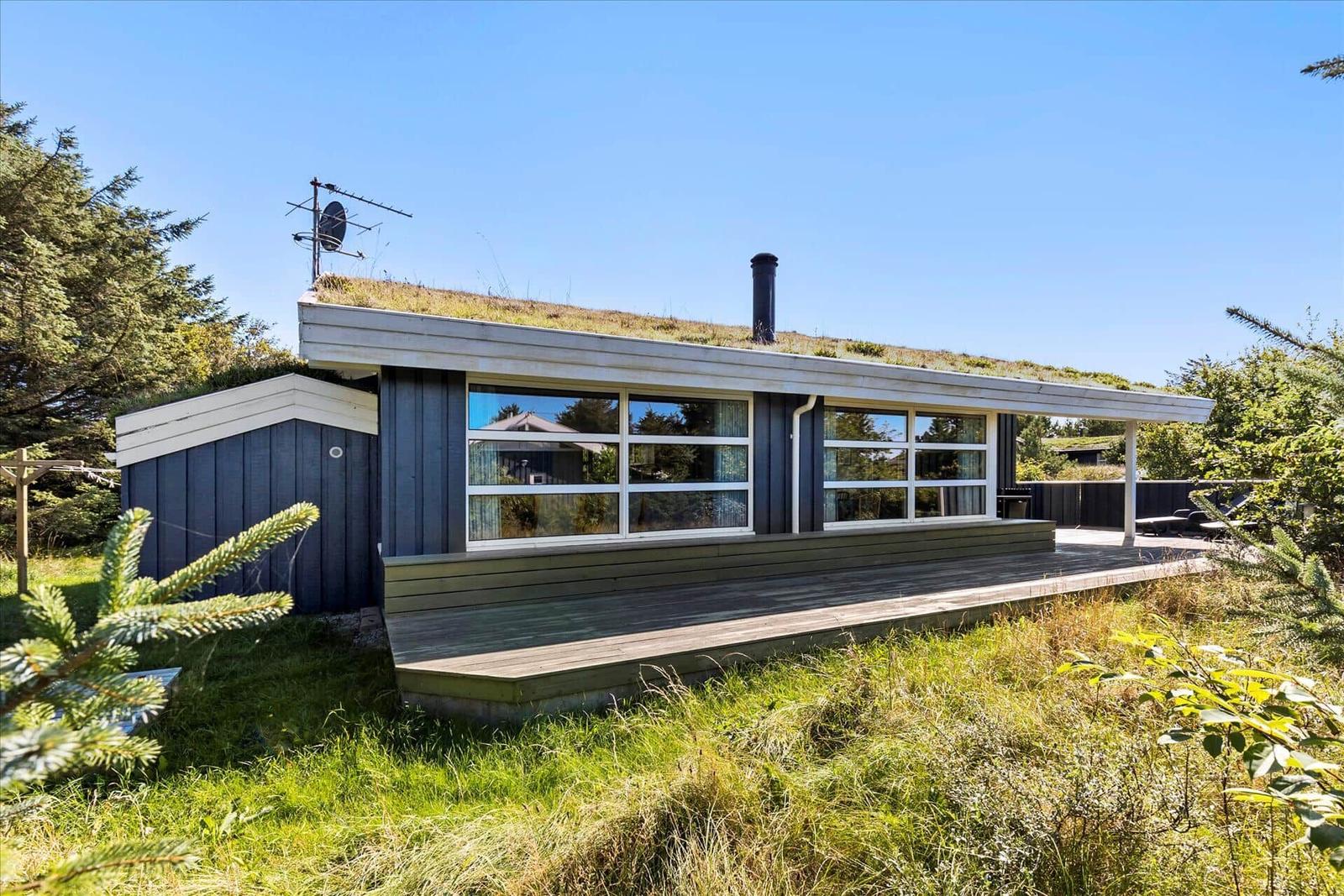 Modern cabin with grass roof, wooden terrace, and large windows in green surroundings.