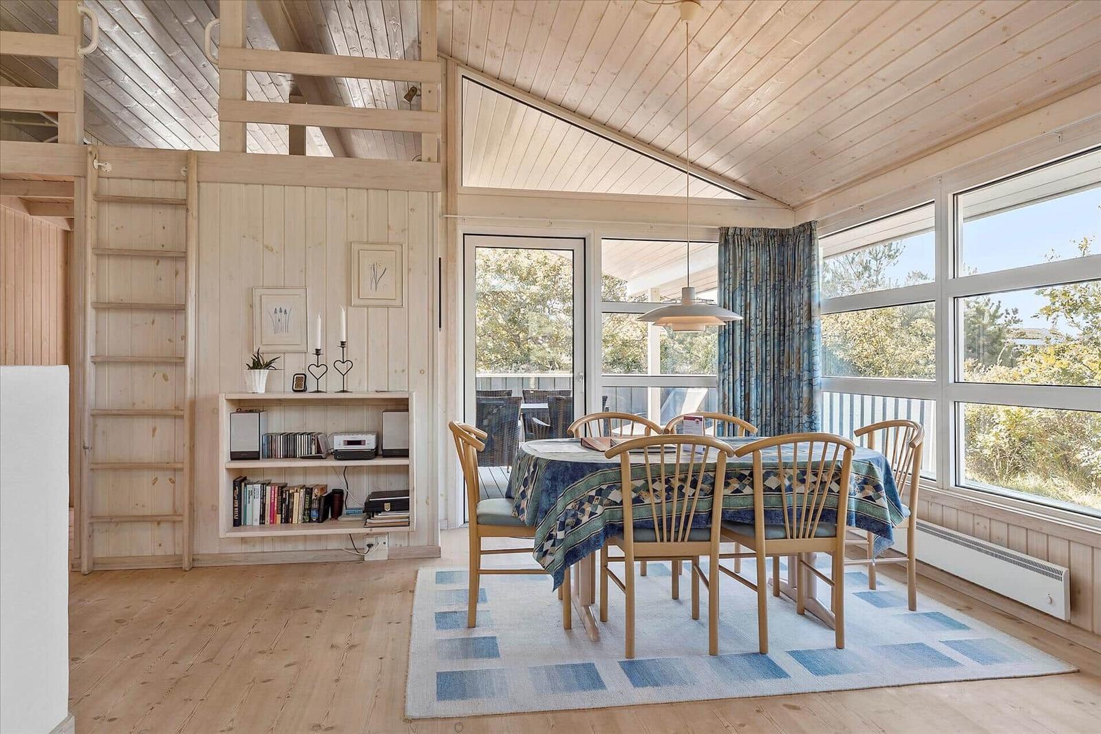 Dining area with table, chairs, and large windows. Wooden ceiling and walls. Staircase in the background.