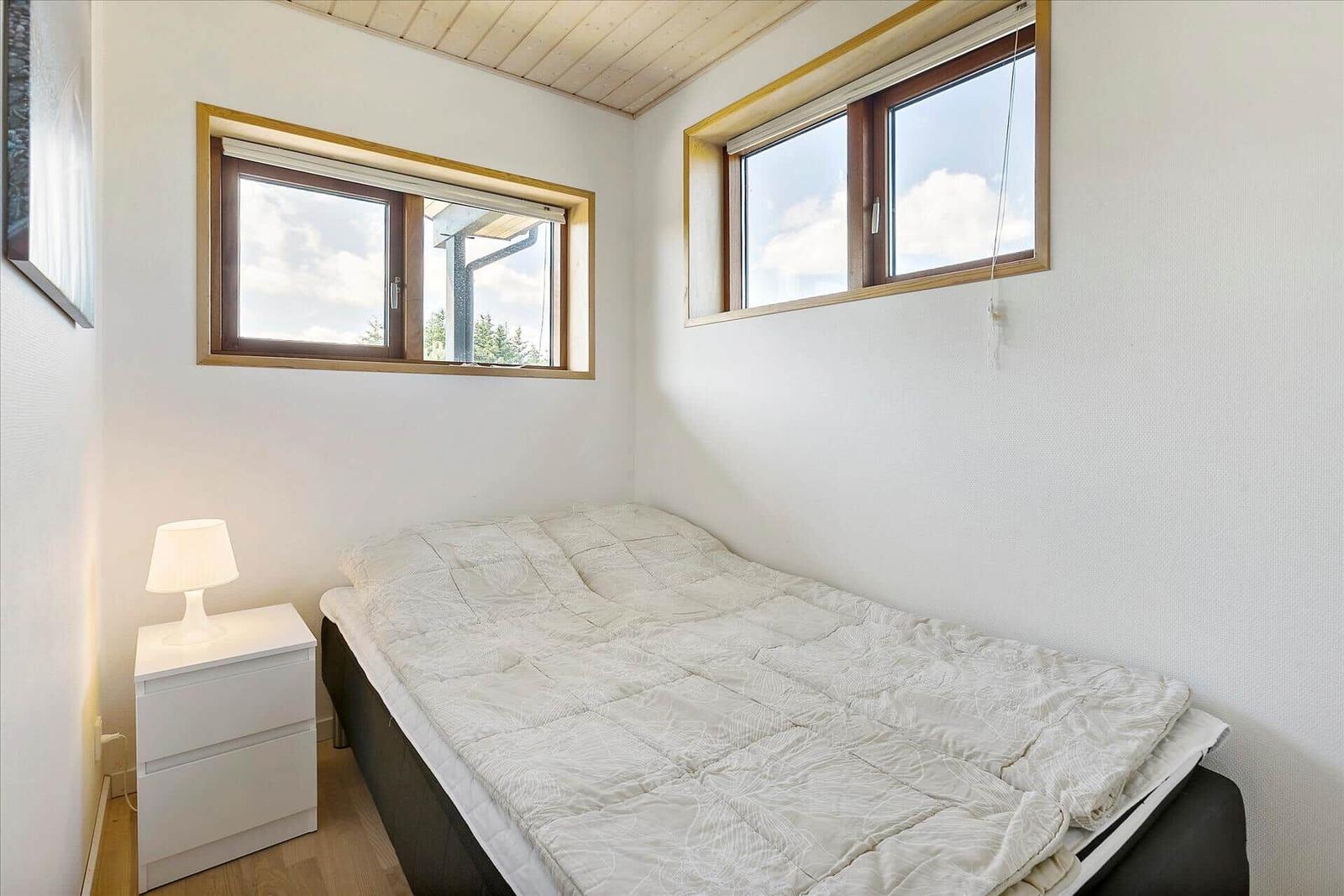 Schlafzimmer mit Bett, Nachttisch und zwei Fenstern. Decke aus Holz.