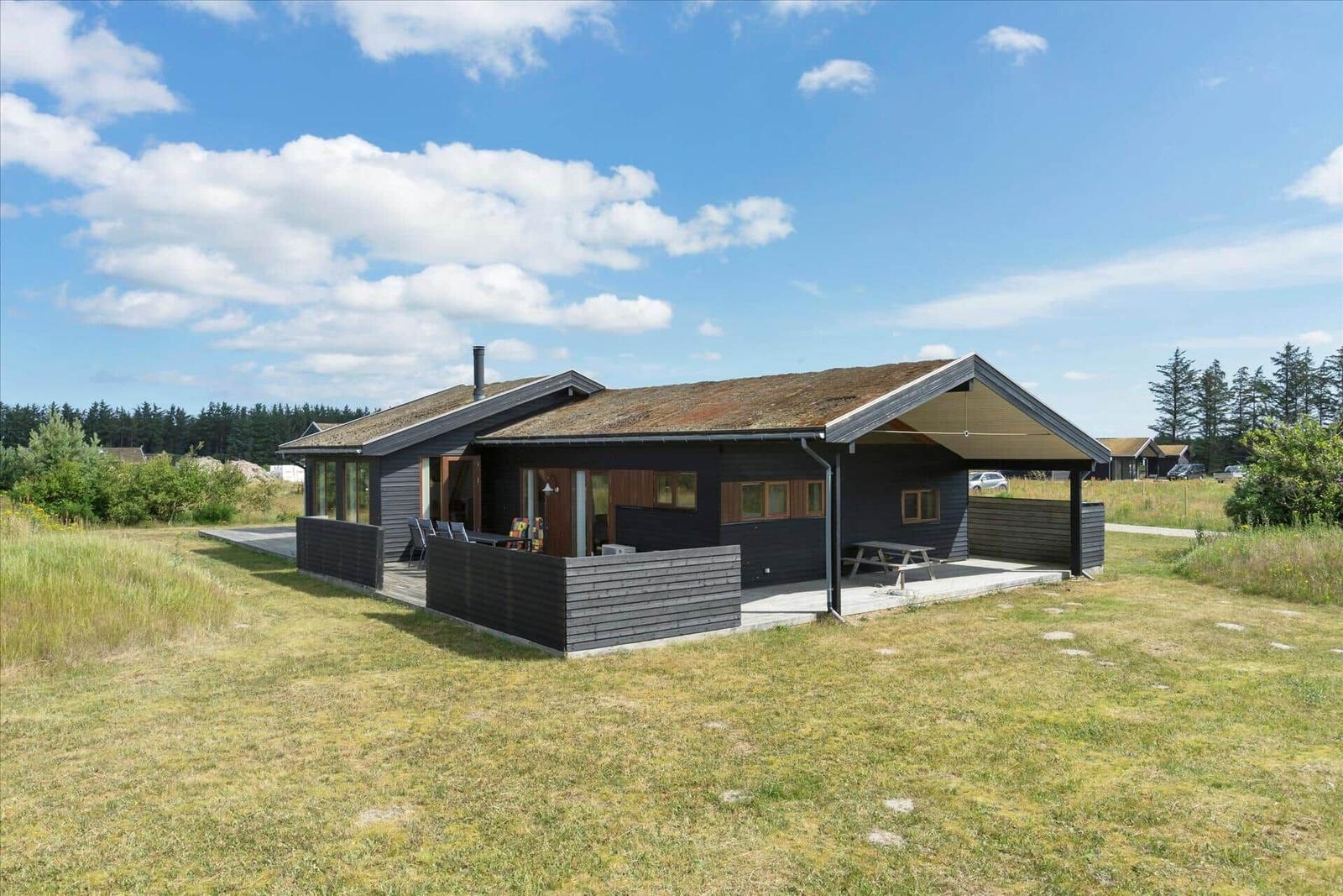 Moderne Holzhütte mit Terrasse und Grillplatz im Grasland unter blauem Himmel.