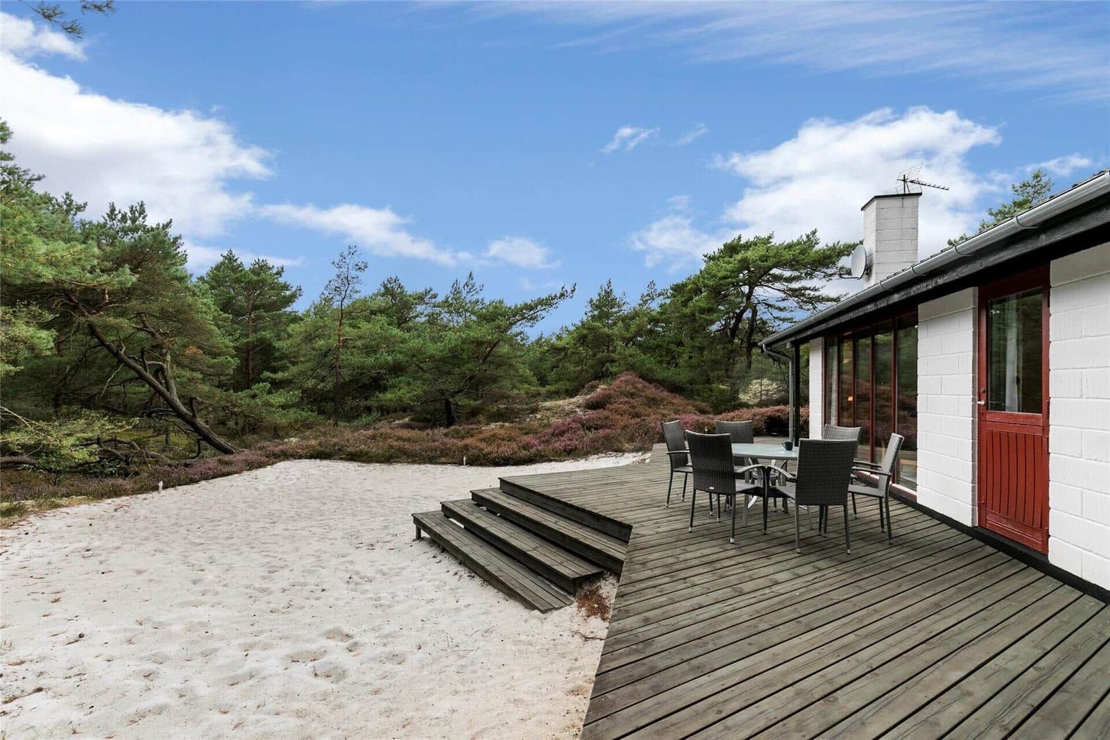 Wohnhaus mit Terrasse, Tisch und Stühlen. Umgeben von Wald und Sandfläche.