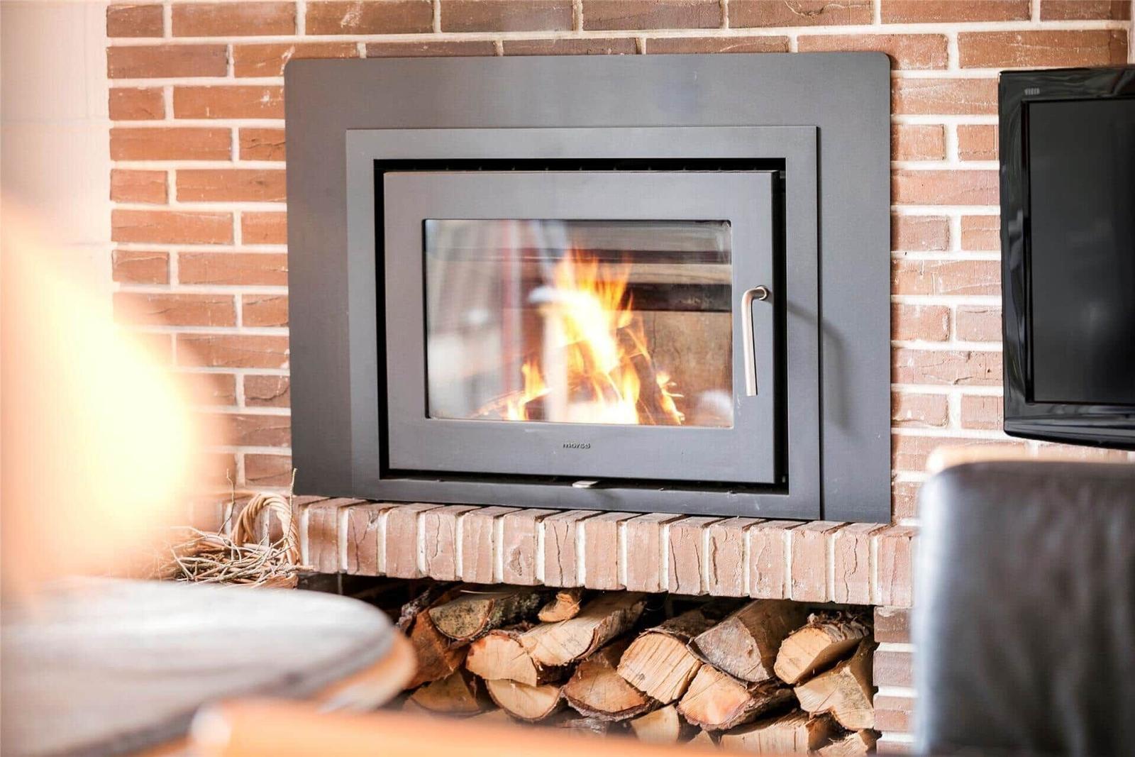 Ein Kamin mit brennendem Holz in einem Raum mit Backsteinwand und Holzstapel darunter.