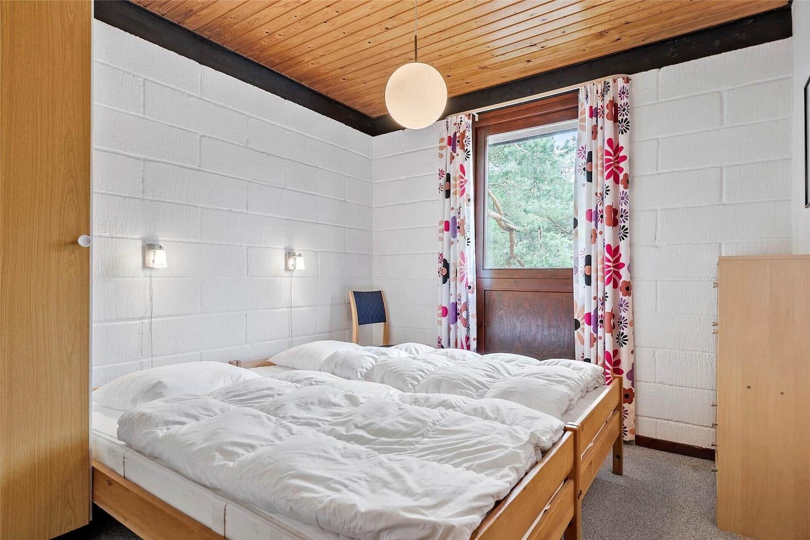 Schlafzimmer mit Doppelbett, Holzboden und Fenster mit floralen Vorhängen.