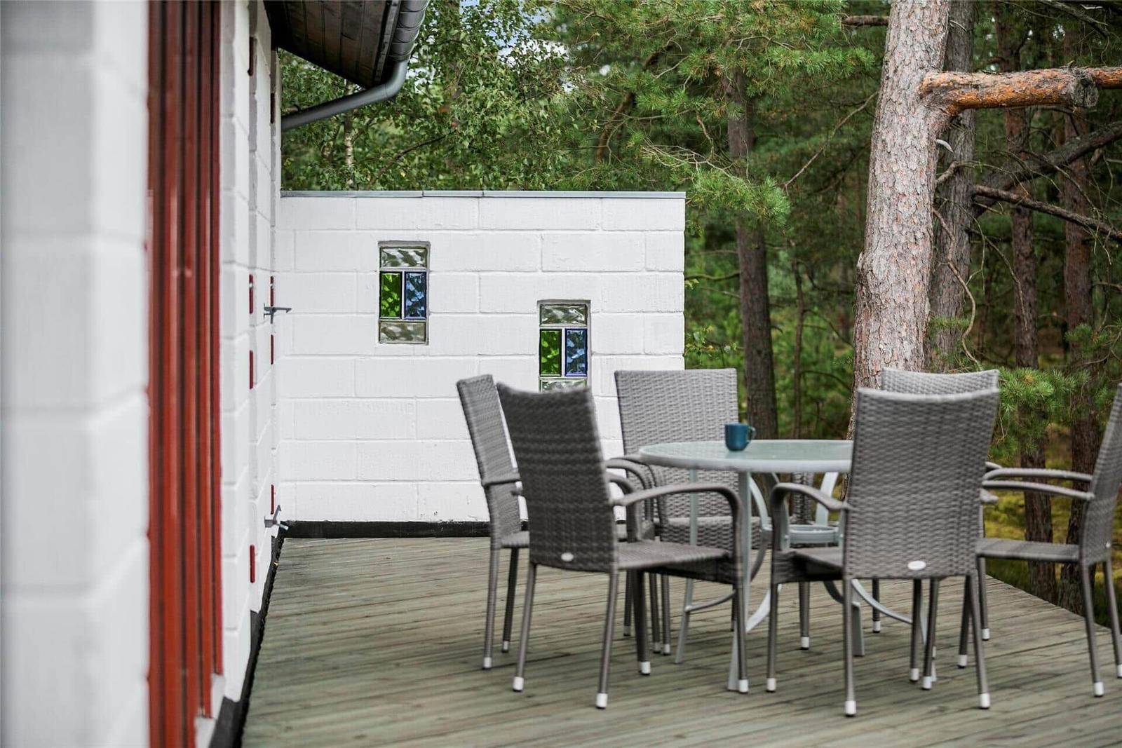 Terrasse mit Tisch und Stühlen, weißer Blockbau mit farbigen Fenstern, umgeben von Bäumen.