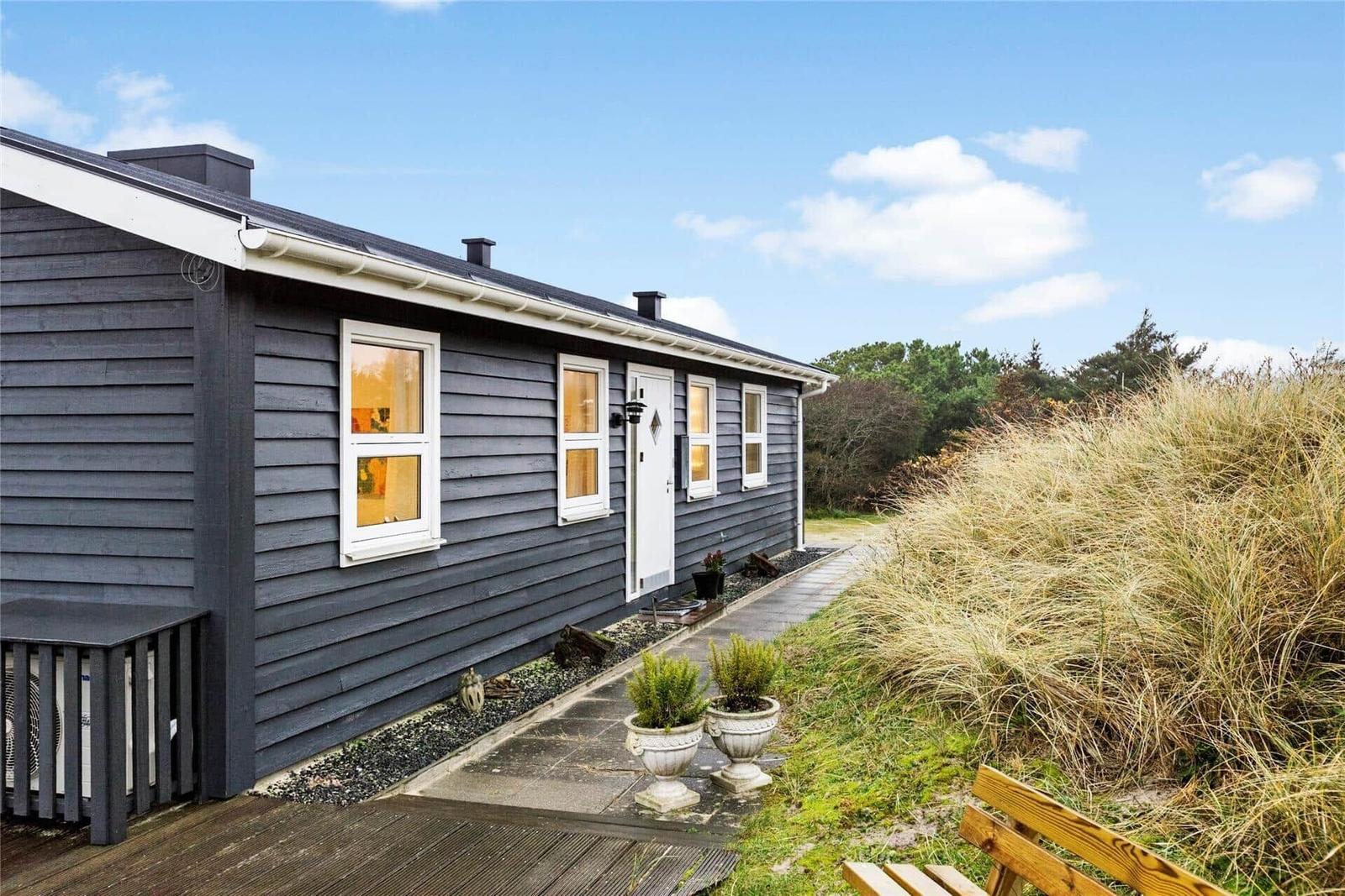 Ein graues Haus mit weißen Fenstern und einer Holzterrasse. Ein Weg führt zu der Tür.