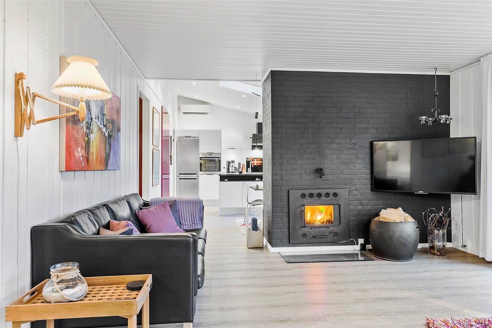 Wohnbereich mit Kamin, Sofa und Fernseher. Küche im Hintergrund sichtbar.