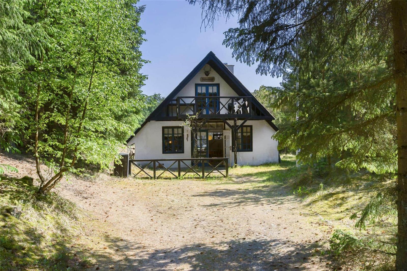 Ein weißes Haus mit dunklem Dach steht im Wald. Es hat eine Veranda und Fenster mit dunklen Rahmen.