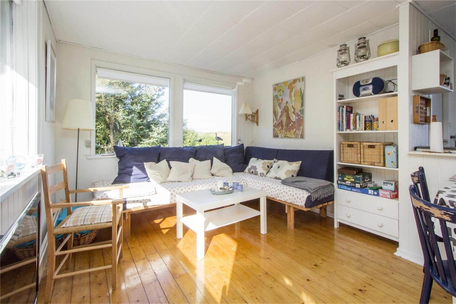 Living room with sofa, table, and bookshelf. Wooden floor and windows with view outside.