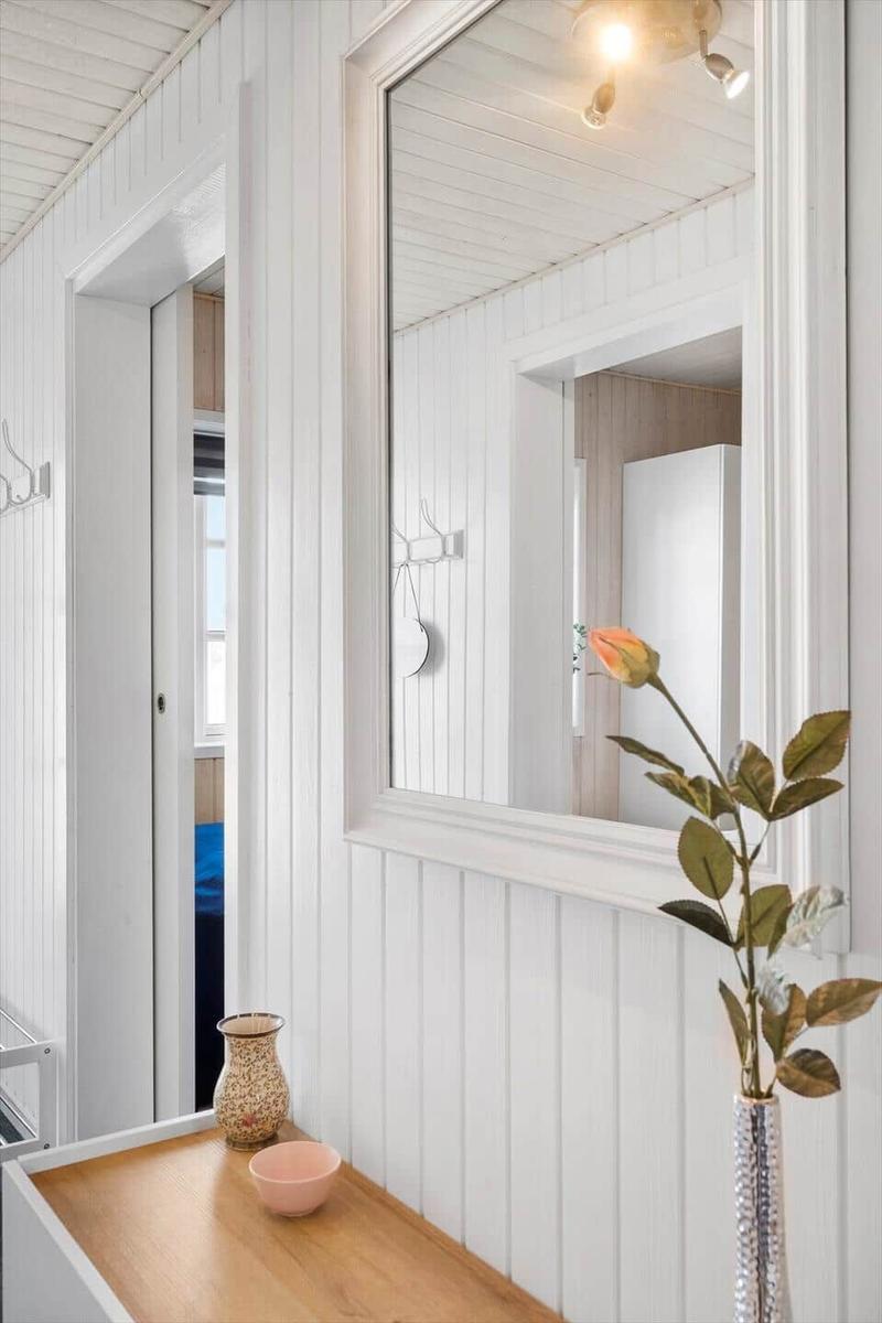 Mirror on white wall, wooden table with vase and flower, doorway to bedroom.