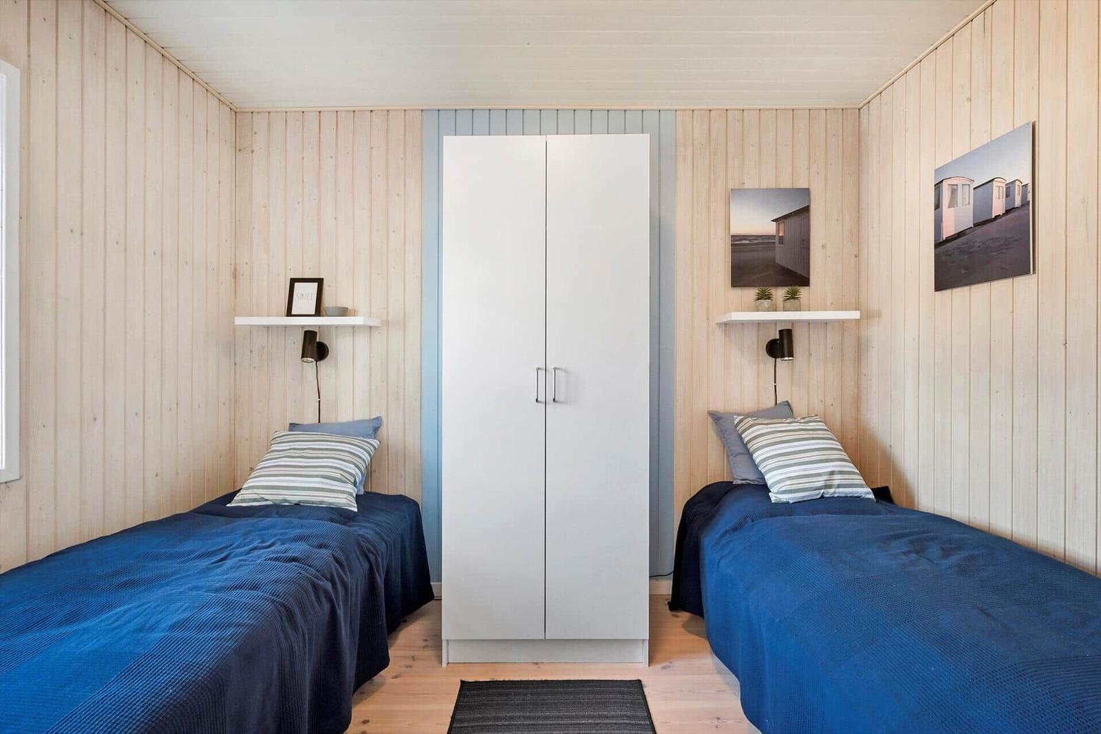 Two beds with blue bedding, white wardrobe, and wood-paneled walls.