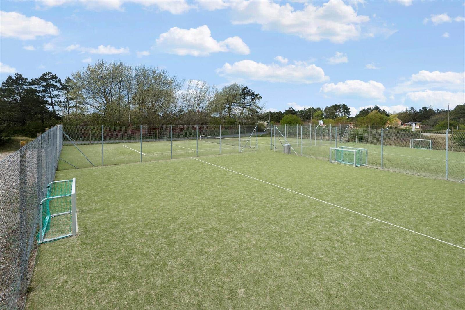 Green sports field with soccer goals and tennis net behind a fence.