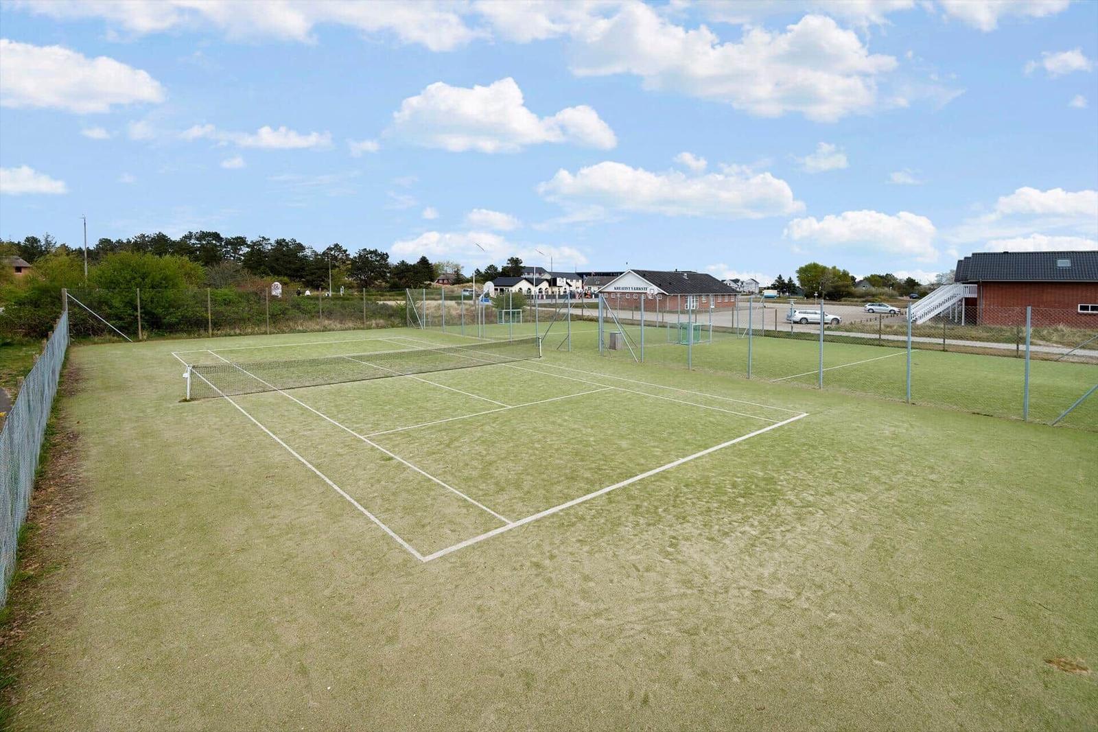 Green tennis court with net and fencing. Buildings and trees are visible in the background.