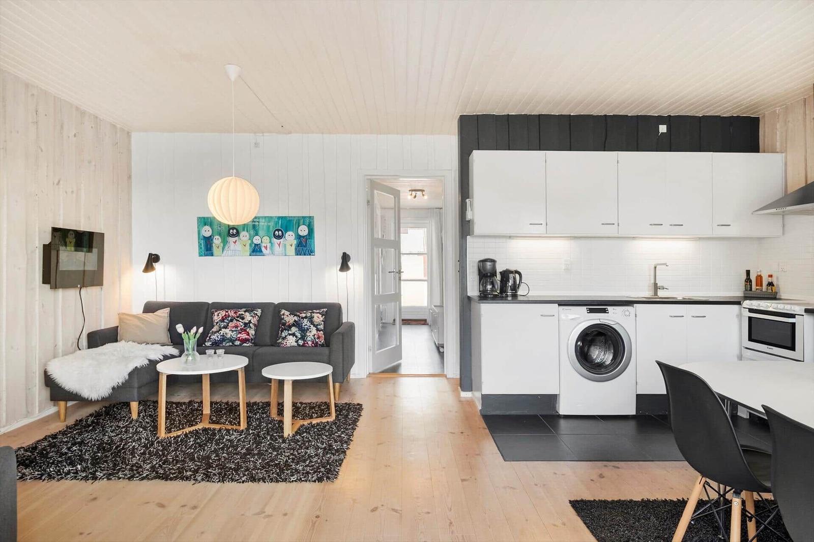 Living area with sofa, tables, and kitchen. White cabinets, washing machine, and black tiles.