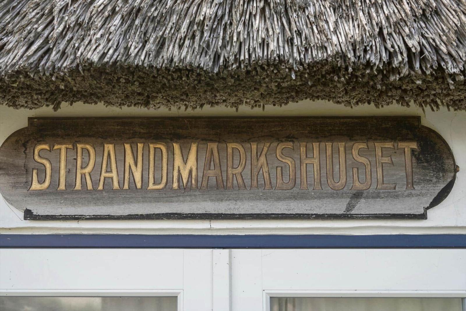 Holzschild mit der Aufschrift STRANDMARKSHUSET unter einem Strohdach.