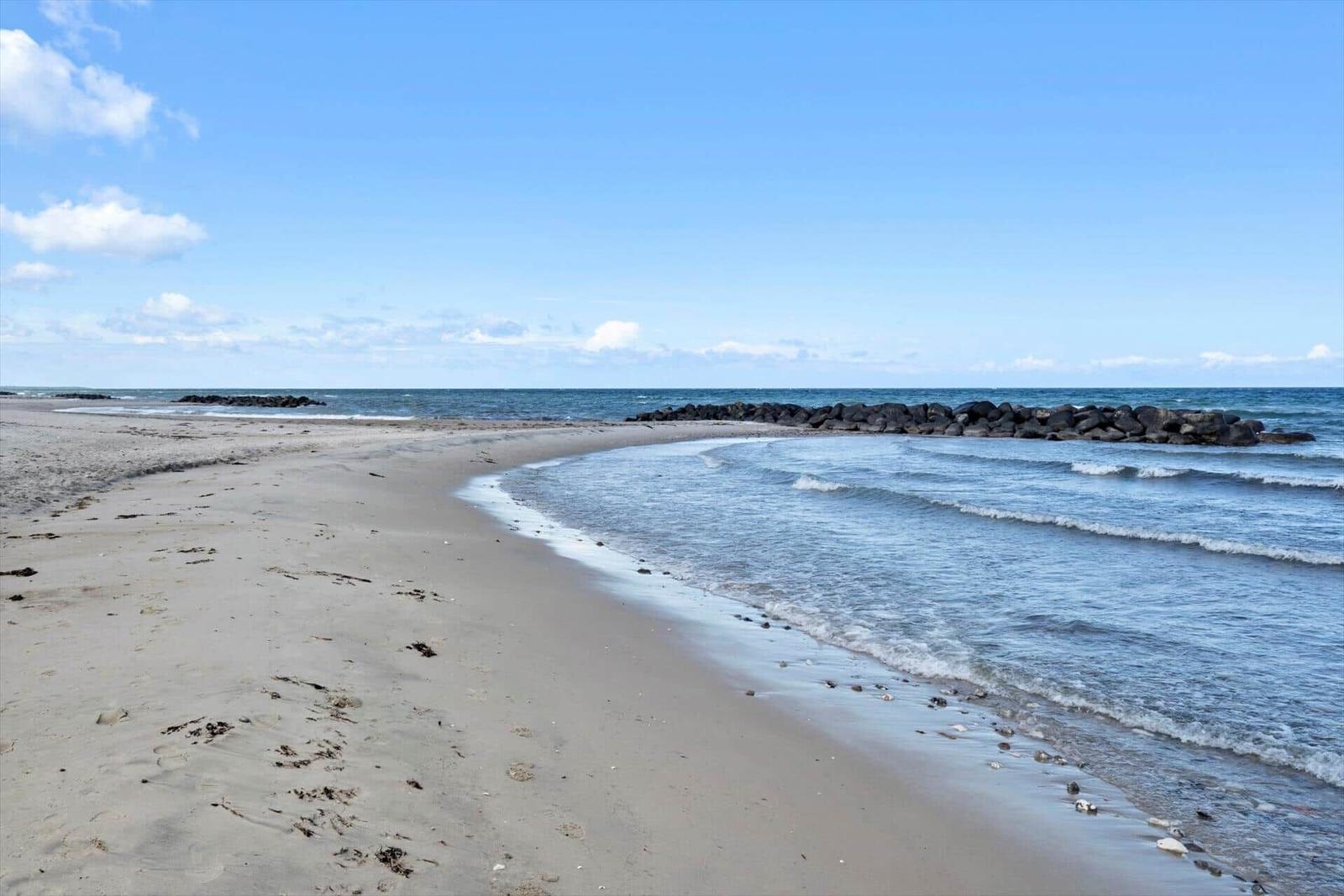 Strand mit Sand und sanften Wellen, Steine am Ufer, klarer Himmel mit wenigen Wolken.