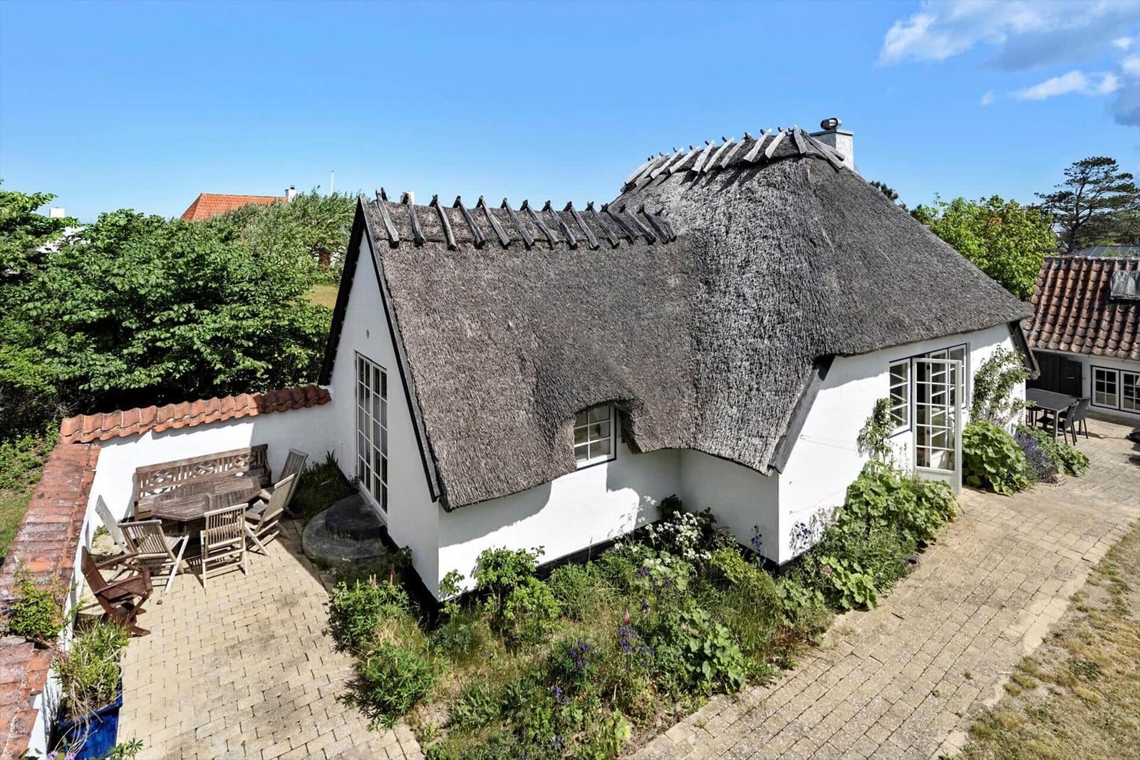 Haus mit Strohdach, Terrasse und Garten. Weiße Wände, Holzmöbel und Pflanzen.