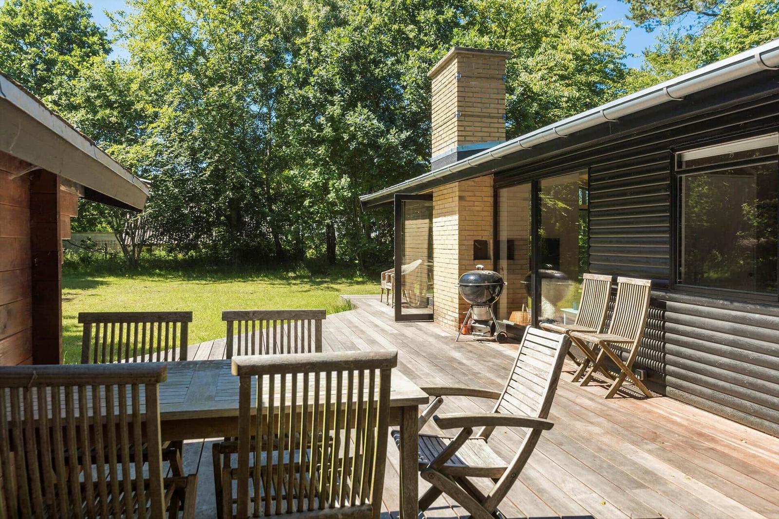 Terrasse mit Holztisch, Stühlen und Grill. Hintergrund: grüner Rasen und Bäume.