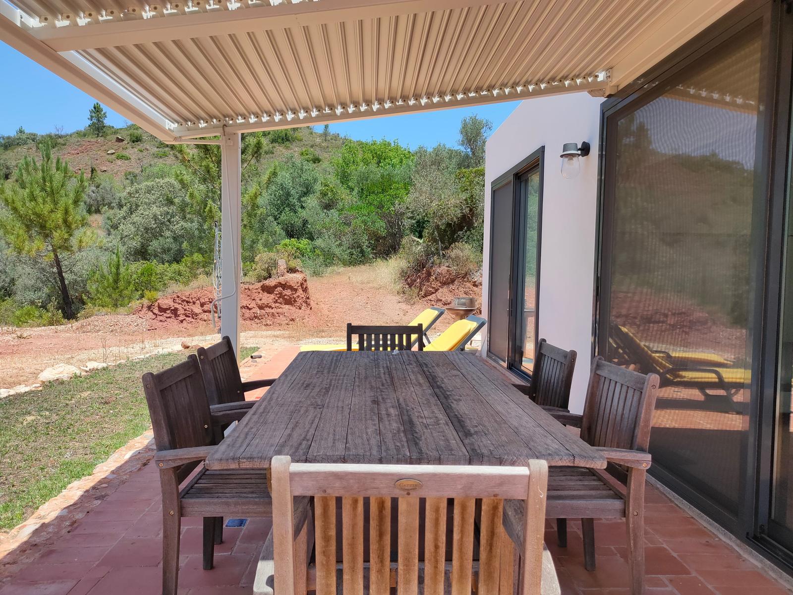 Terrasse mit Holztisch und Stühlen, Blick auf grüne Hügel.