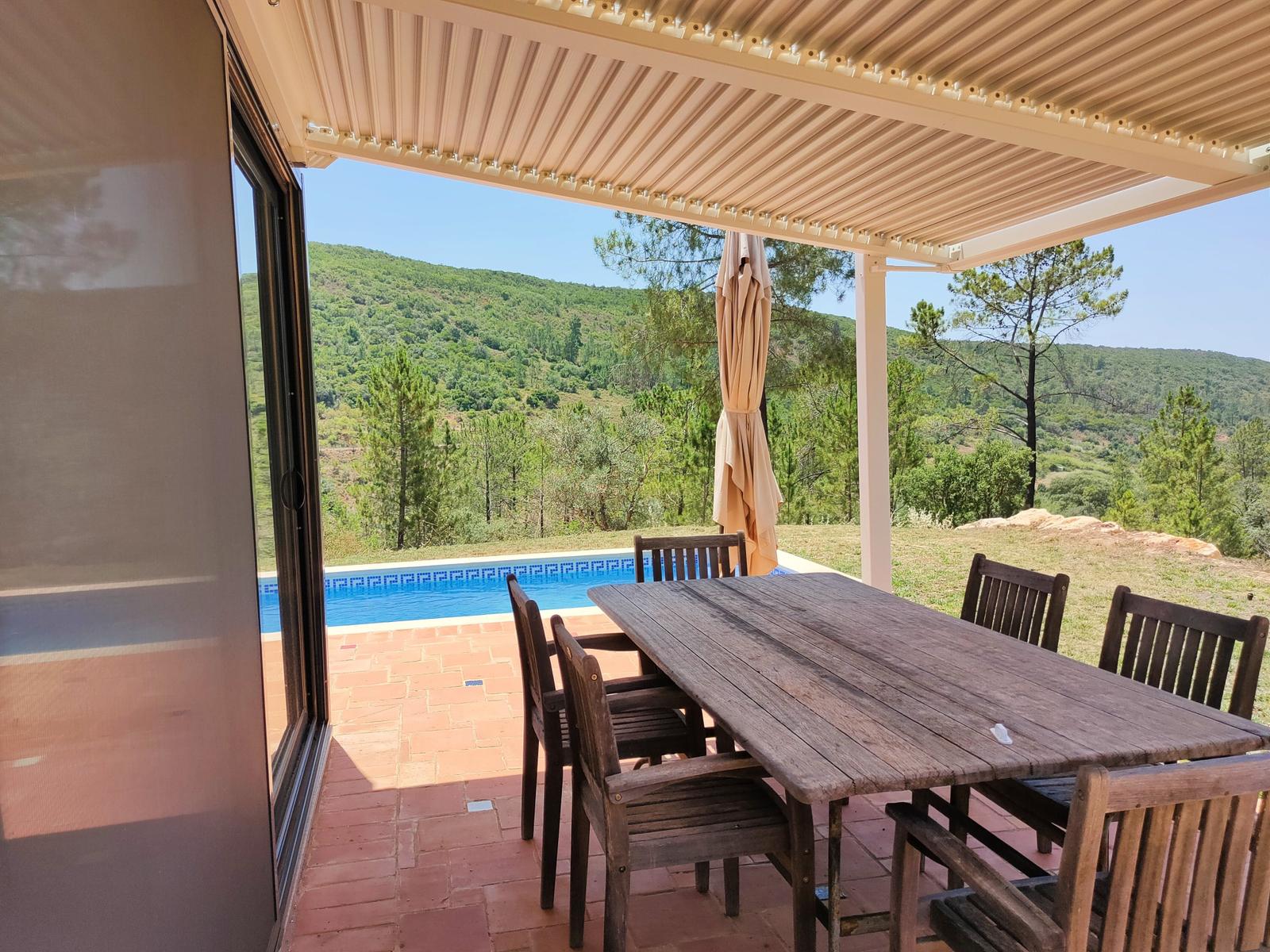 Terrasse mit Holztisch und Stühlen, Blick auf Pool und grüne Hügel.