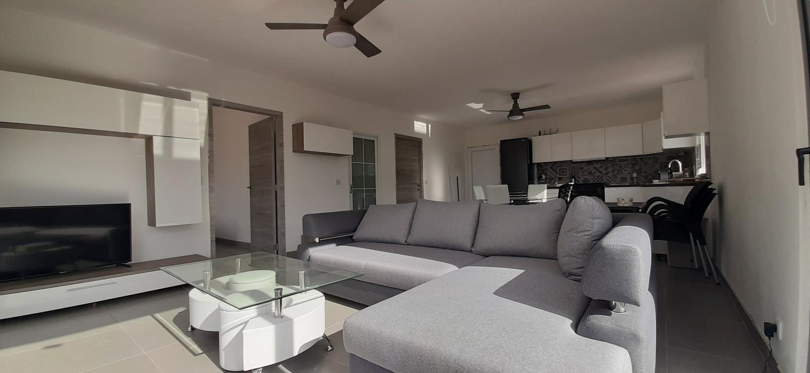 Wohnzimmer mit Sofa, Glas-Tisch und TV. Küche mit Küchenschränken und Arbeitsplatte.