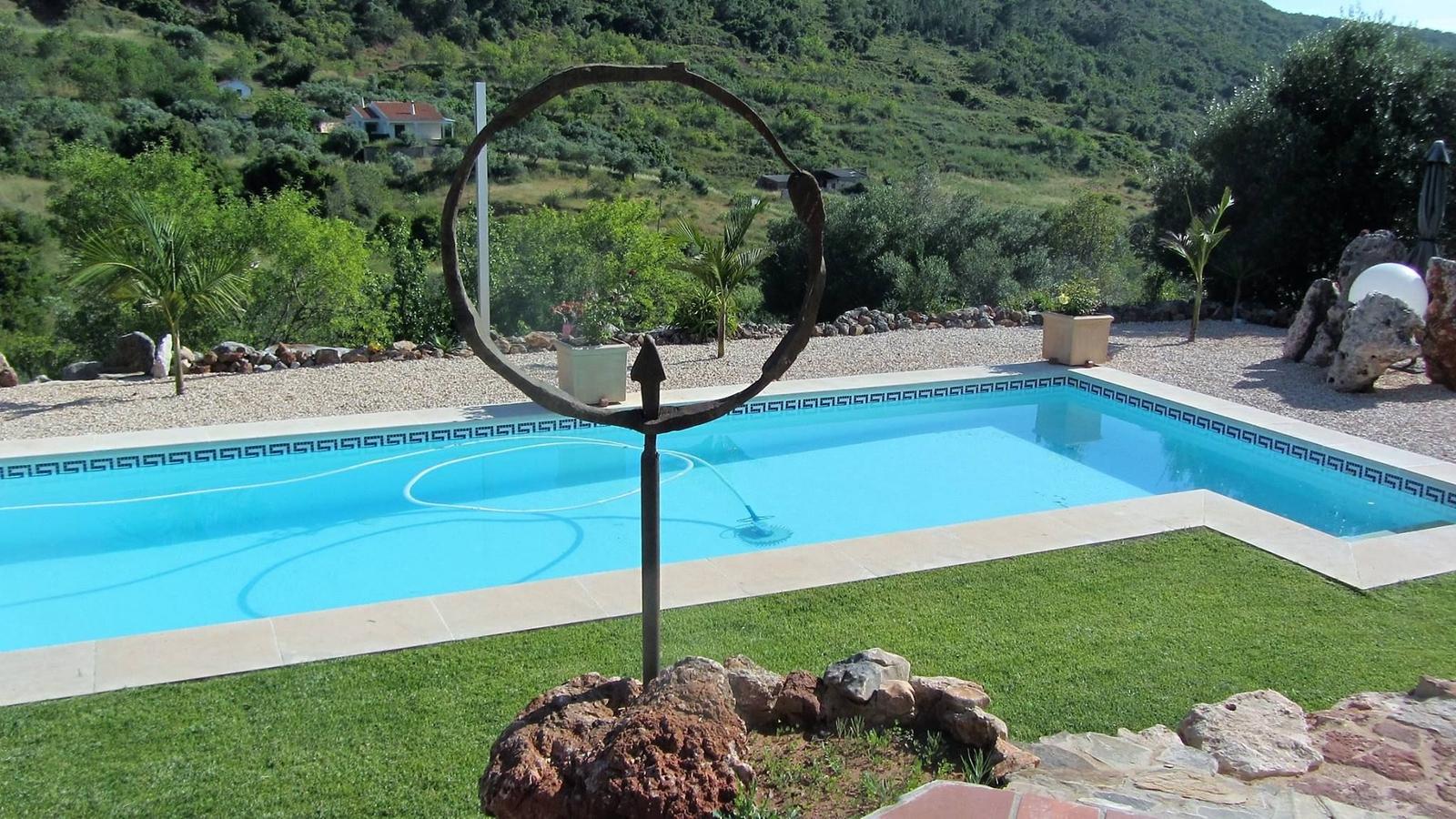 Pool mit Steinumrandung und Skulptur im Vordergrund. Grüne Landschaft im Hintergrund.