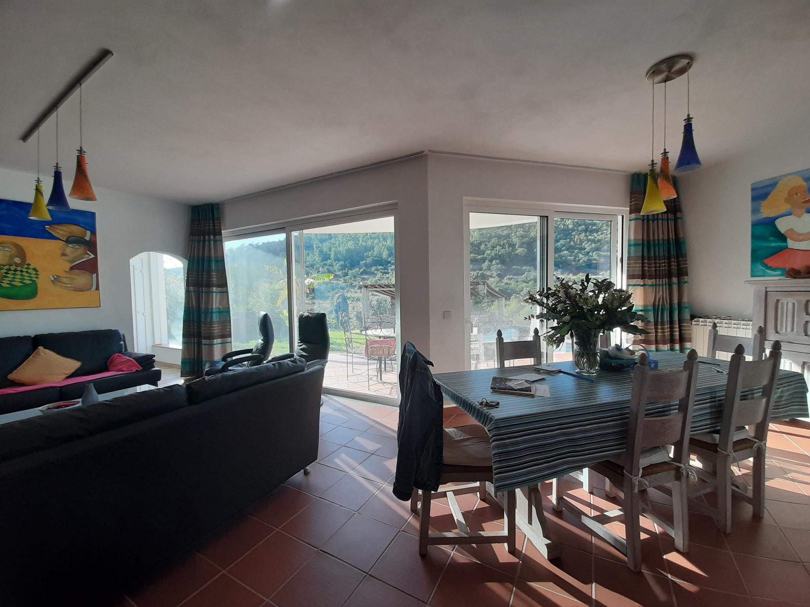 Wohn- und Essbereich mit Blick auf Garten und Hügel. Tisch, Stühle, Sofa und Fenster sind sichtbar.