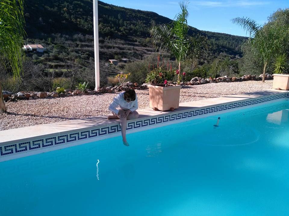 Person sitzt am Poolrand mit Blick auf Hügel und Palmen.