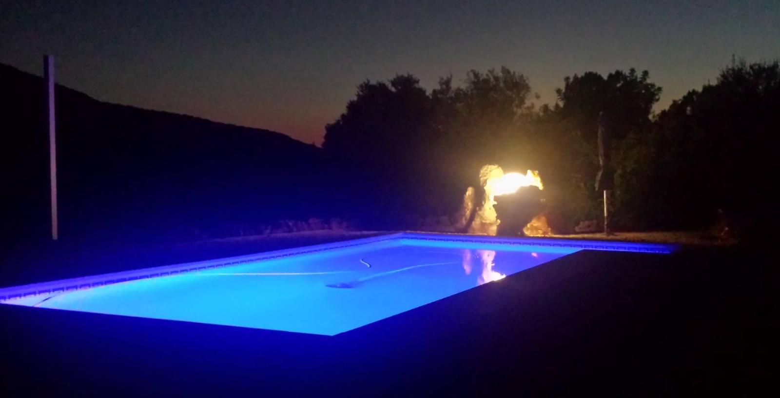 Beleuchteter Pool bei Nacht mit Feuerfontäne im Hintergrund.