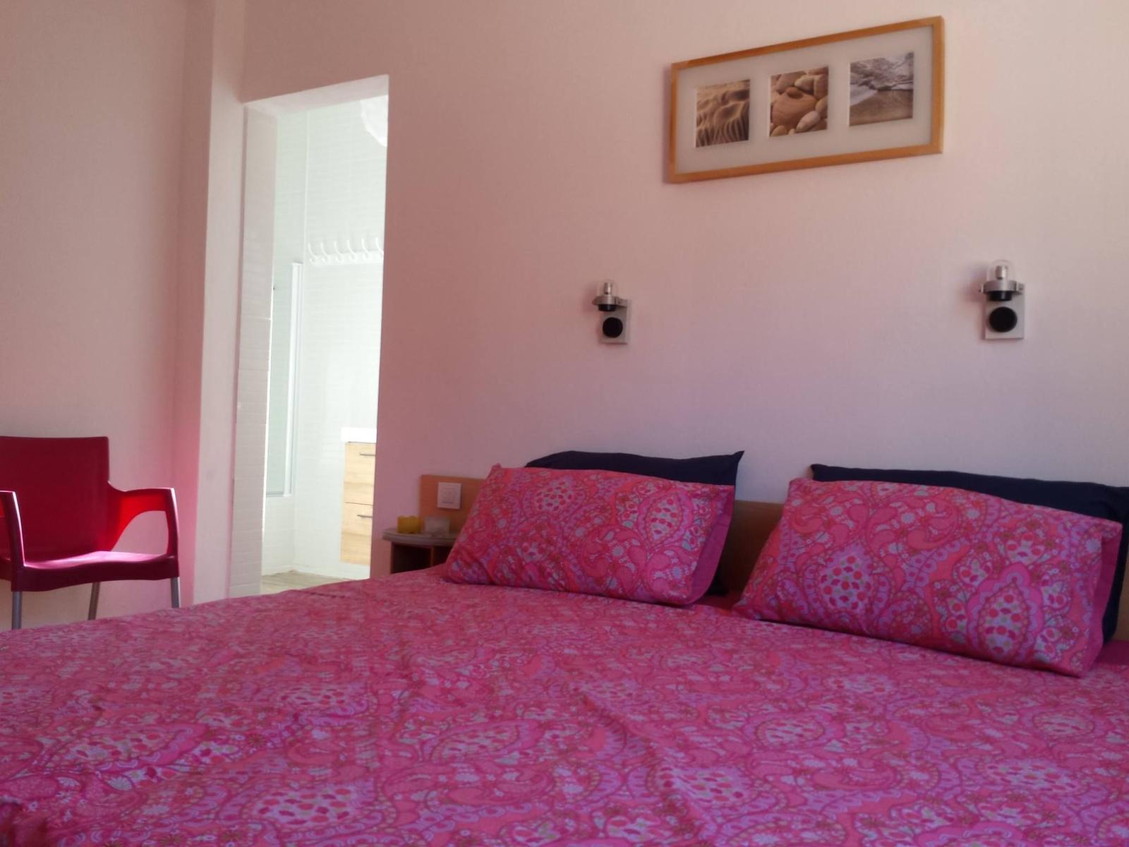 Schlafzimmer mit Bett, Kissen und Wandbild. Rote Sitzgelegenheit und angrenzender Flur.