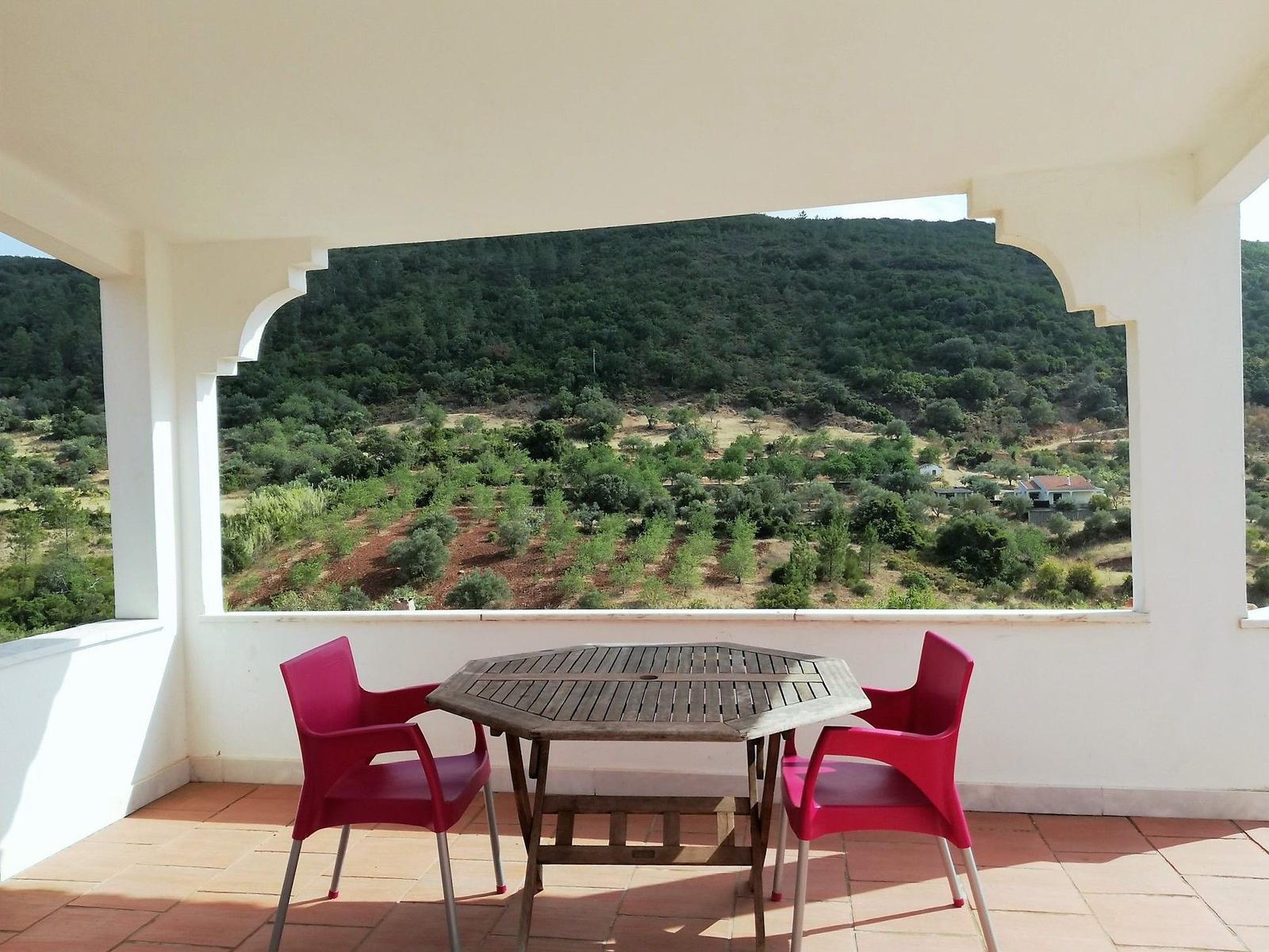 Terrasse mit Tisch und Stühlen, Blick auf grüne Hügellandschaft.