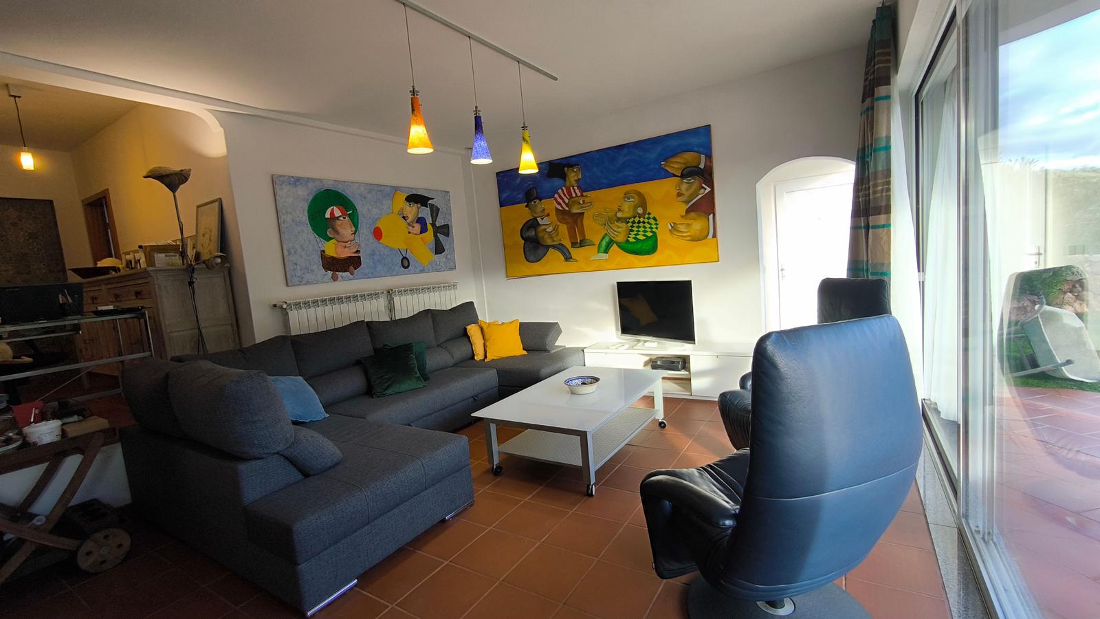 Wohnzimmer mit Sofa, Couchtisch und großen Fenstern. Wandbilder und Hängelampen sichtbar.
