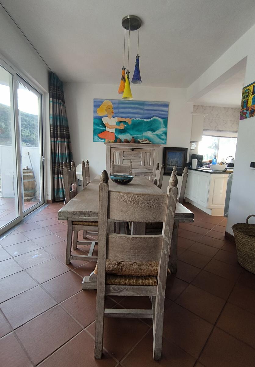 Esszimmer mit Holztisch, Stühlen und Wandbild. Küche im Hintergrund. Terrasse über Glastür.