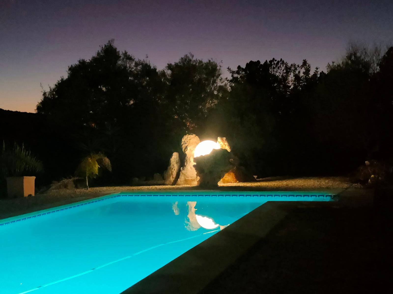 Beleuchteter Pool bei Nacht mit Steinformation und Bäumen im Hintergrund.