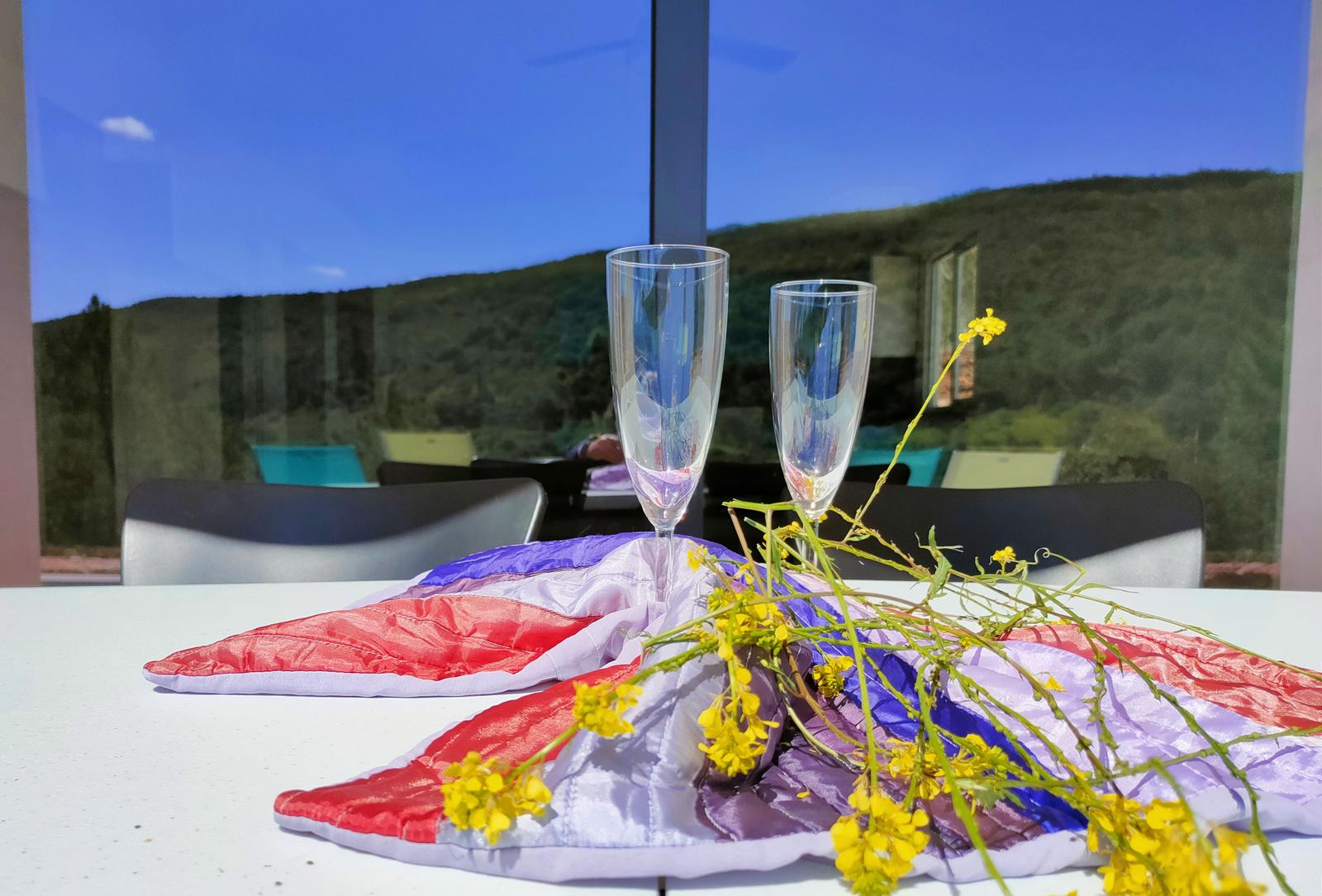 Tisch mit zwei Gläsern, Blumen und buntem Tuch vor großem Fenster.