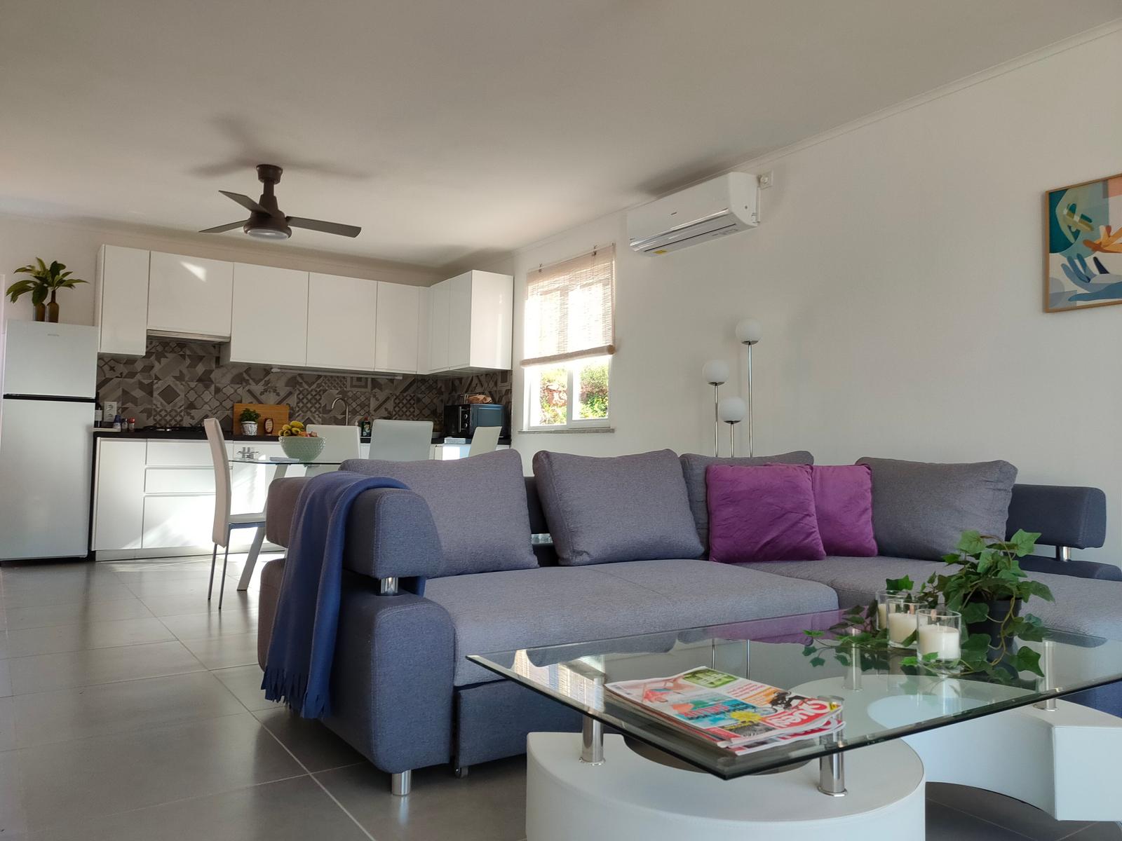 Wohnzimmer mit Sofa, Glas-Tisch und Küchenbereich im Hintergrund.