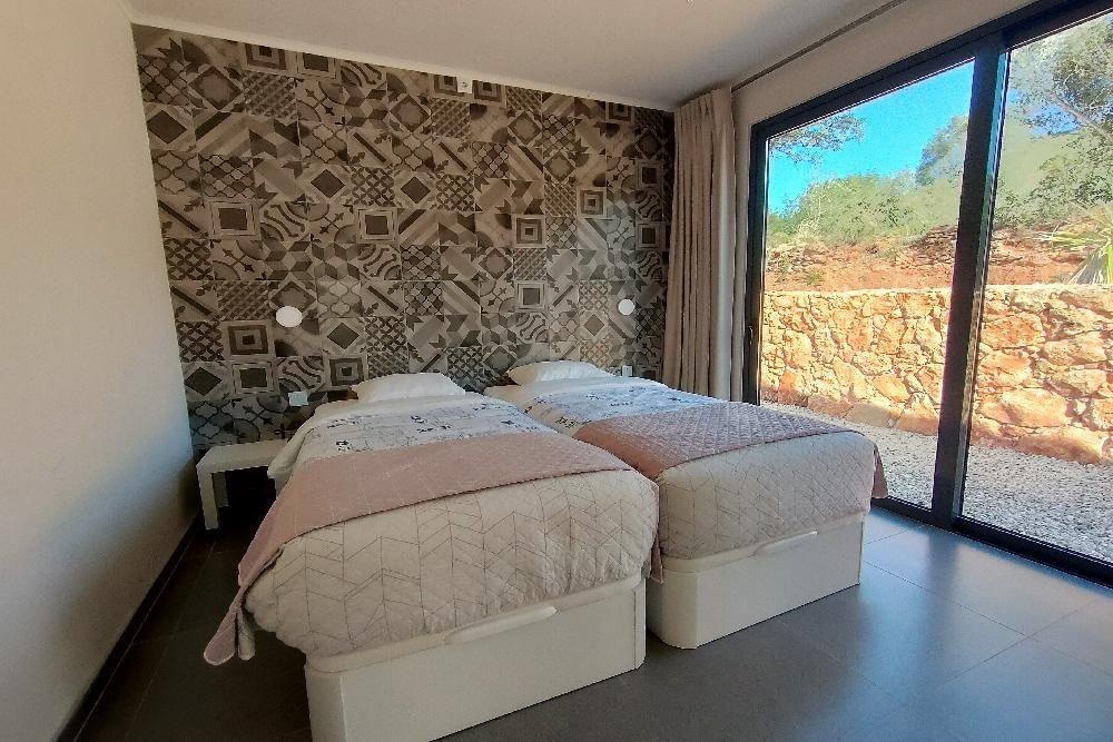 Zwei Betten mit gemusterten Bettwäsche und einem Mosaikwandpaneel. Große Glastür mit Blick auf Steinmauer.