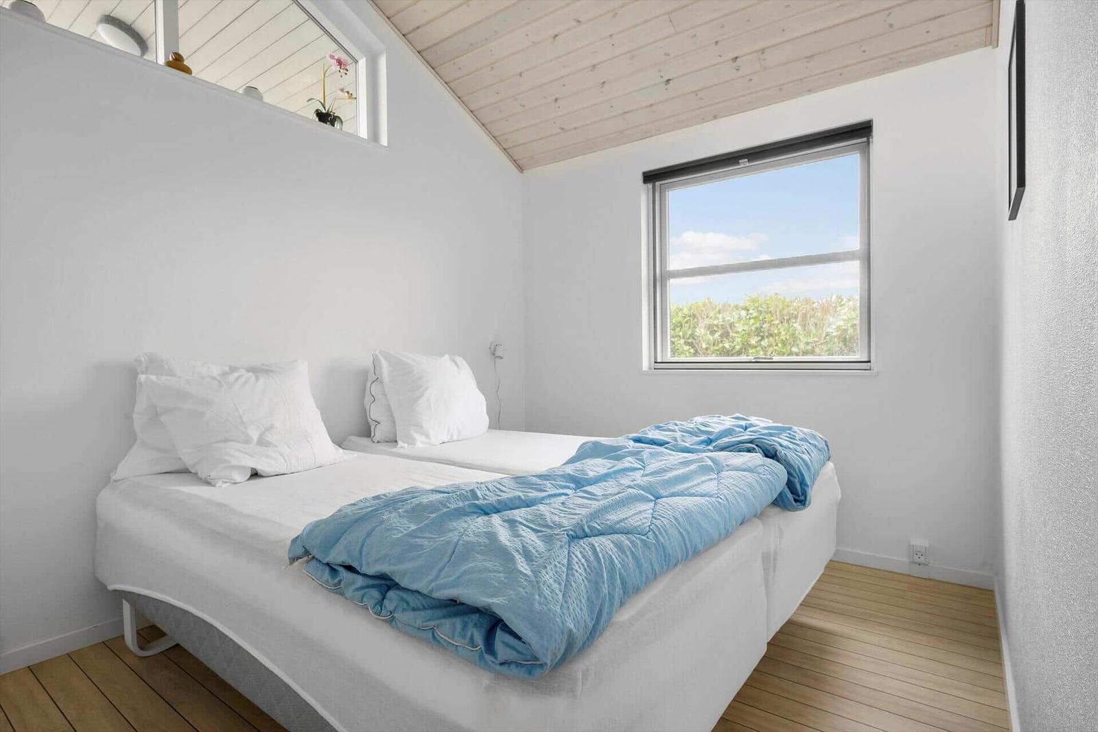 Schlafzimmer mit Doppelbett, blauer Decke und Fensterblick auf Grünflächen.