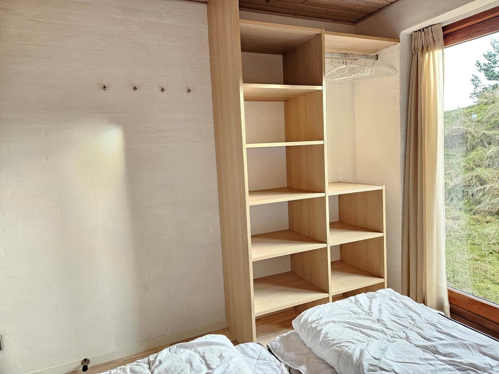 Schlafzimmer mit Holzregal, Bett und Fenster mit Blick auf Grünfläche.