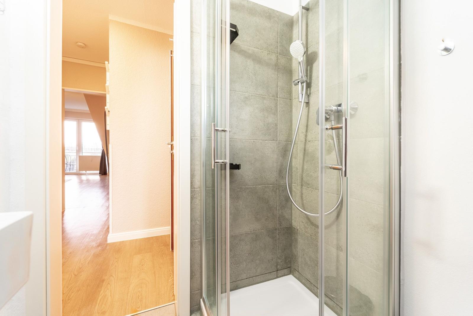 Dusche mit Glaswand und grauen Fliesen. Blick in Wohnbereich mit Holzboden.