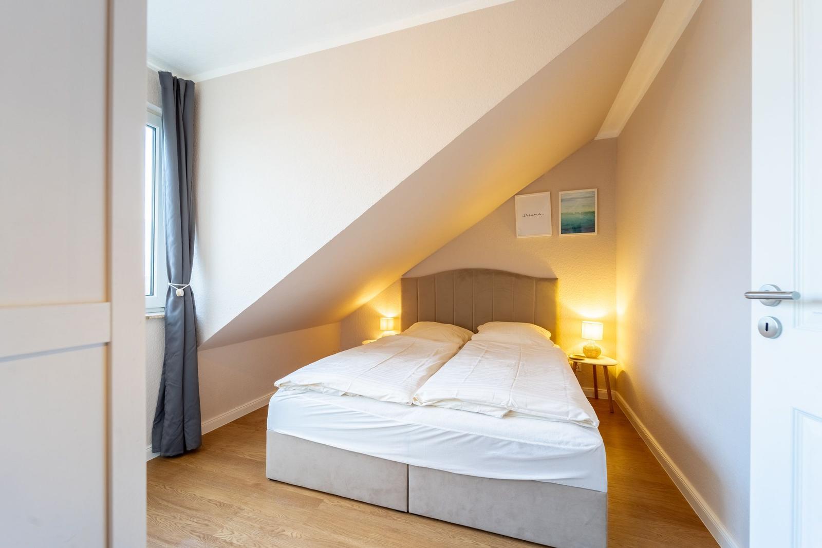 Schlafzimmer mit Dachschrägen, Bett, Nachttisch und Fenster mit Vorhang.