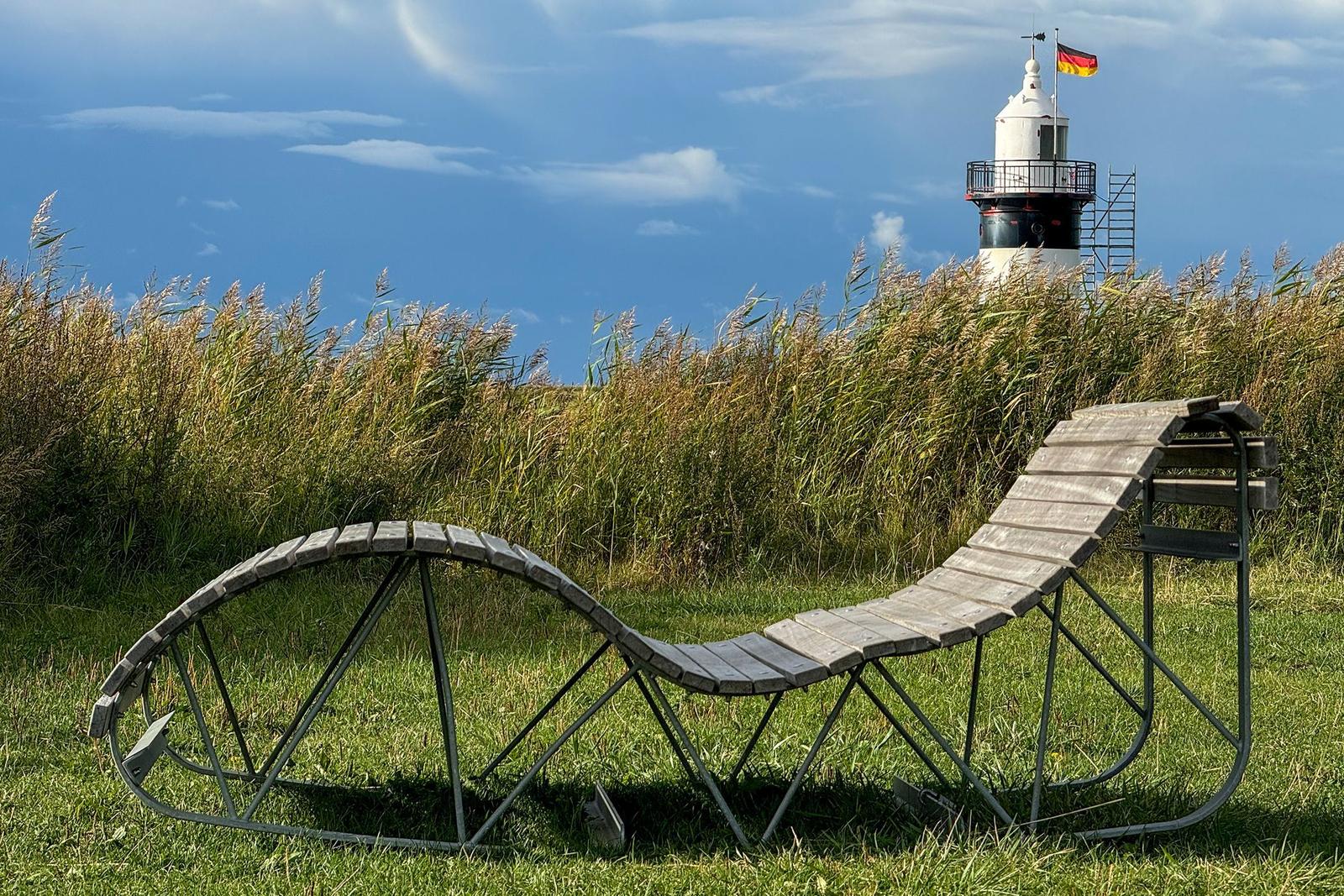 Holzliege im Gras vor einem Leuchtturm mit deutscher Flagge.