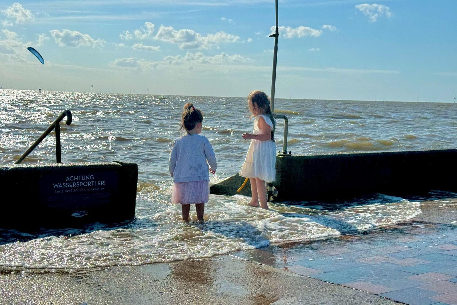 Zwei Kinder stehen am Ufer und spielen im Wasser. Im Hintergrund ist das Meer und ein Kitesurfer zu sehen.