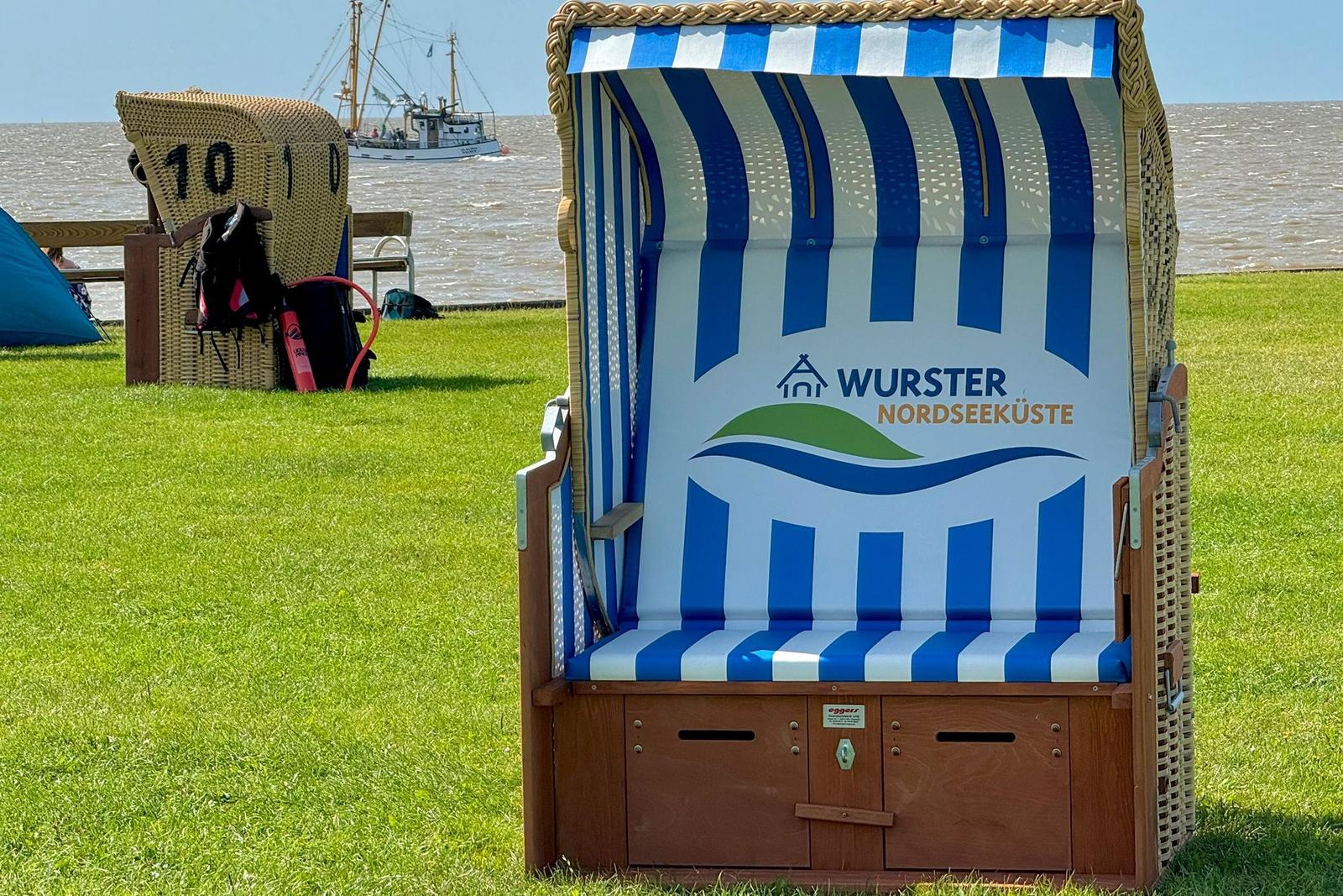 Strandkorb mit blau-weißem Muster und Logo auf grünem Rasen. Im Hintergrund See und Boot.