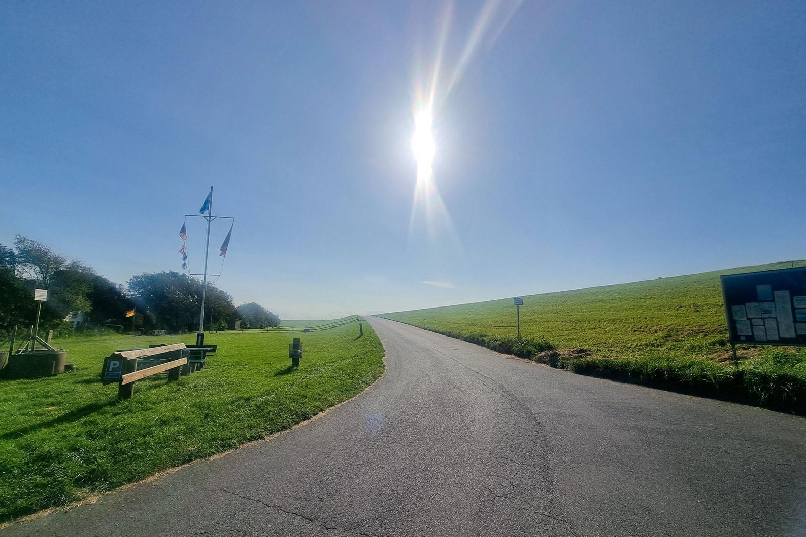 Sonniger Weg durch grünes Feld mit Parkplatz und Flaggenpfosten.