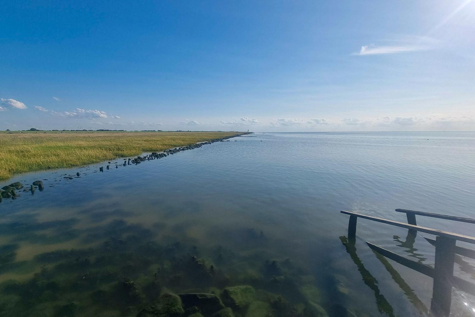 Wasserfläche mit Steinen und Holzbrücke im Vordergrund, Grasland und Windräder im Hintergrund.