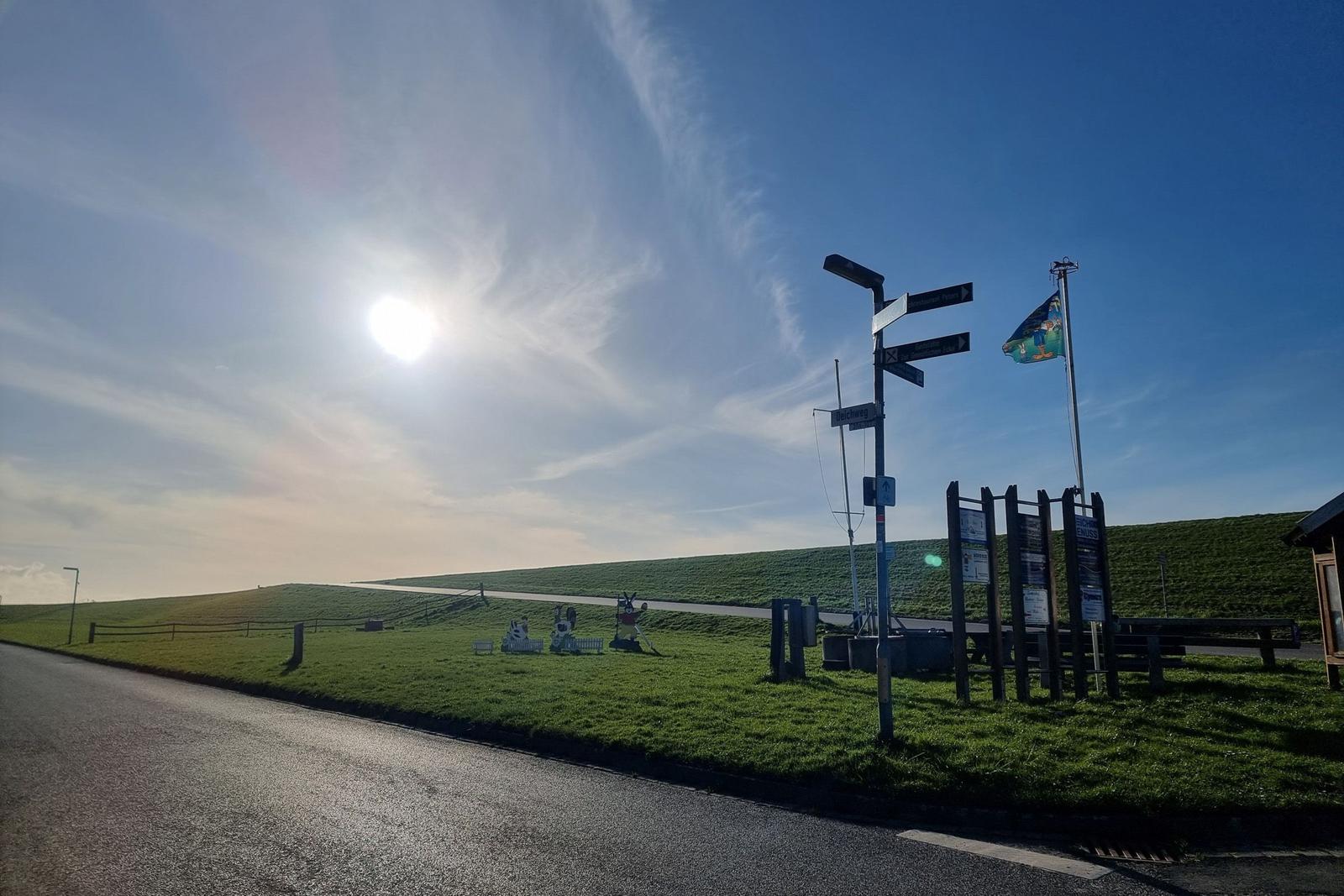 Sonniger Tag mit Wegweiser und Fahnenmast an einer grünen Hangstraße.