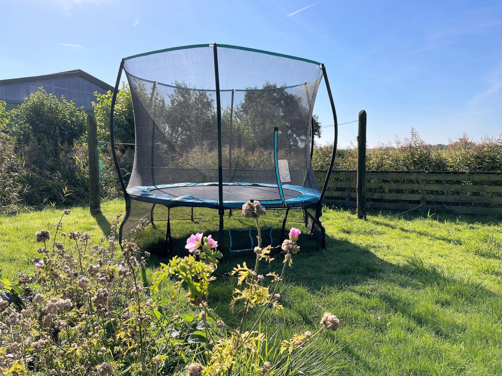 Trampoline with net in green garden with wooden fence and background vegetation.