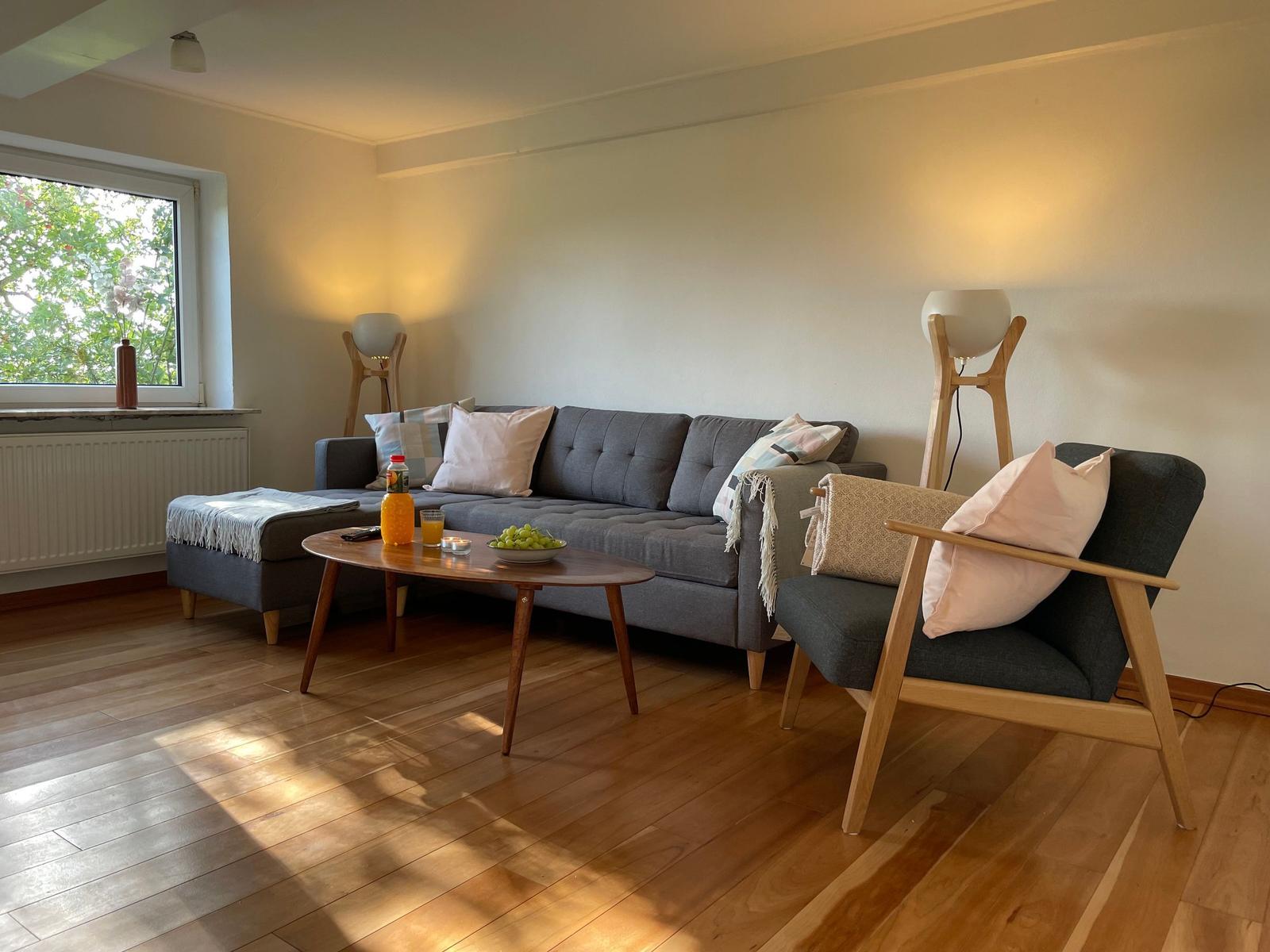 Living room with sofa, armchair, and wooden table. Window with view of trees.